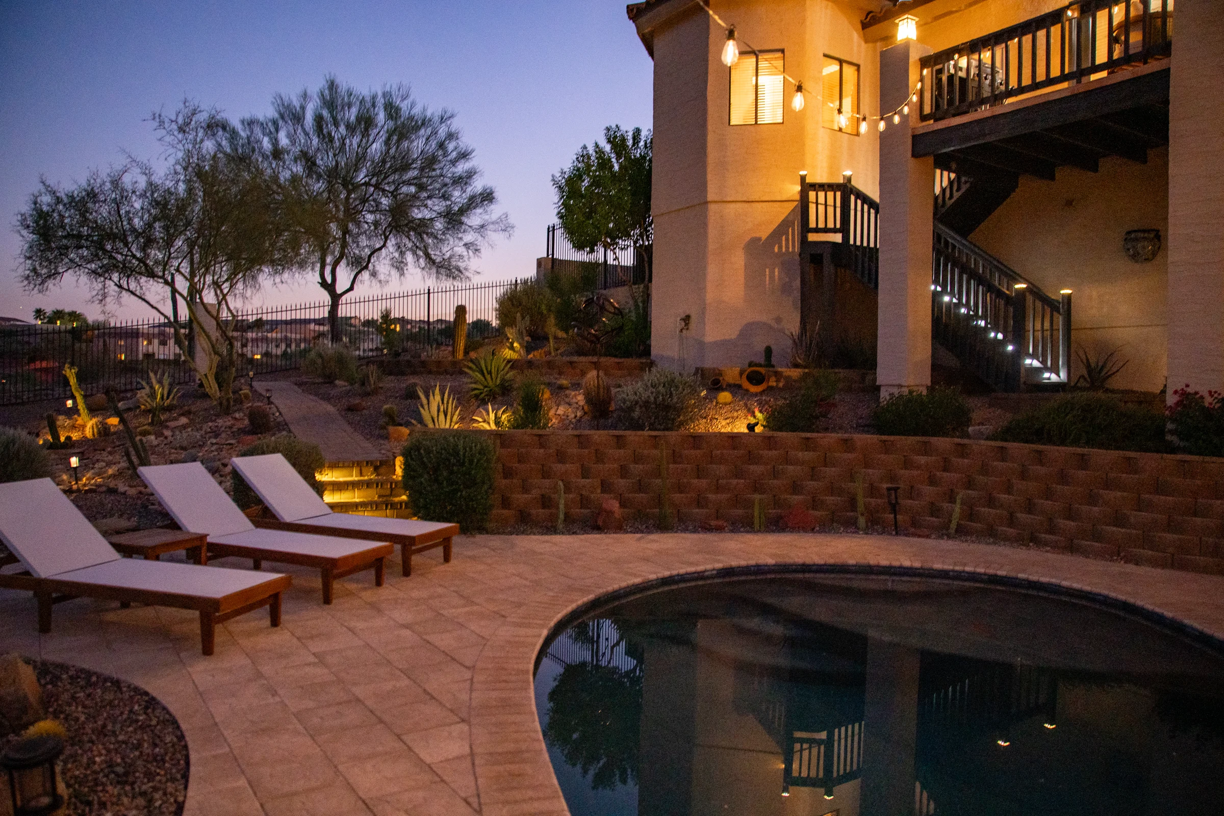 Evening view of a backyard oasis featuring a pool, lounge chairs, illuminated desert landscaping, and a two-story house with string lights. Photographed by Shane Baker Studios.