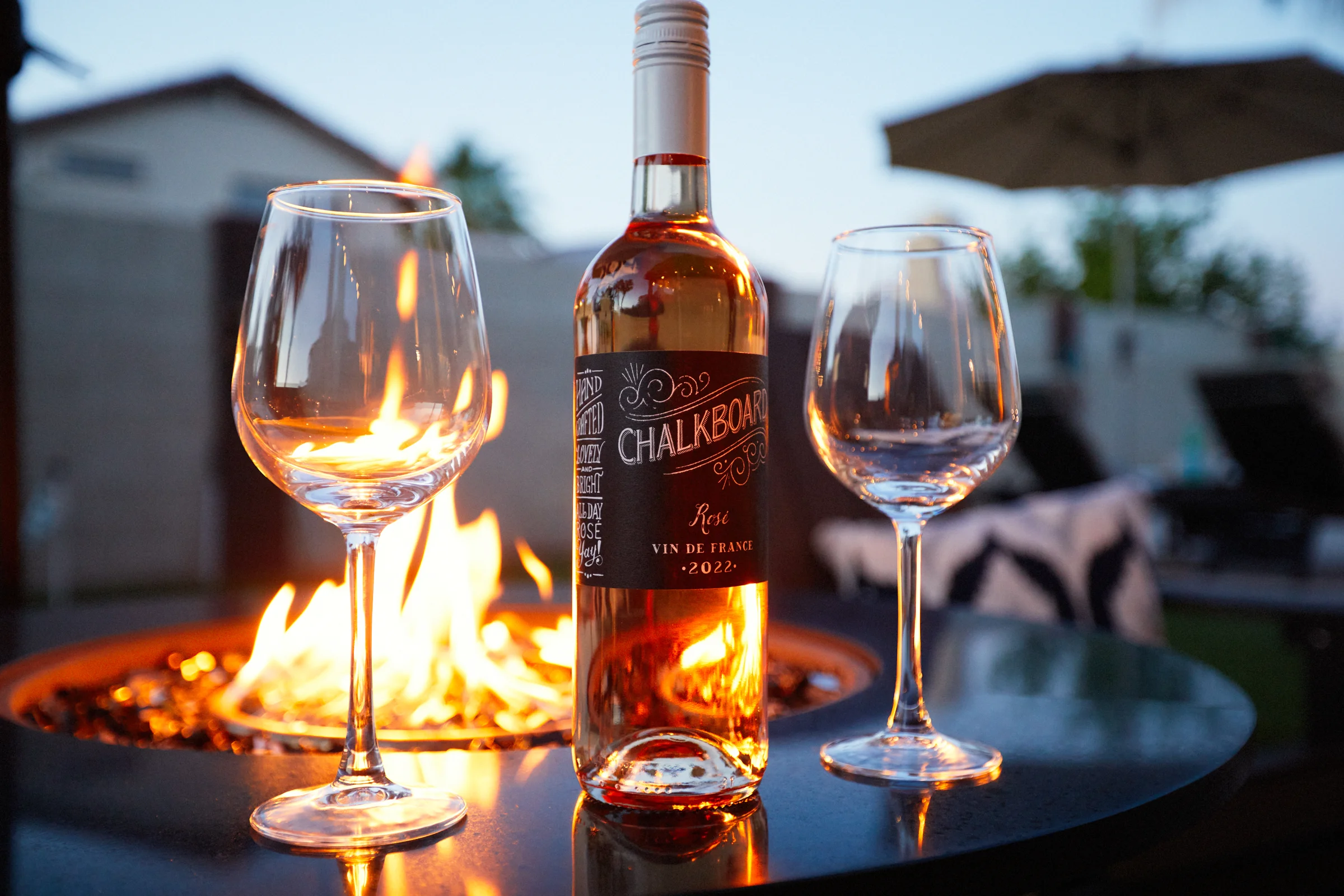 A bottle of Chalkboard Rosé wine with two empty wine glasses, illuminated by the warm glow of a fire pit during twilight. Photographed by Shane Baker Studios.