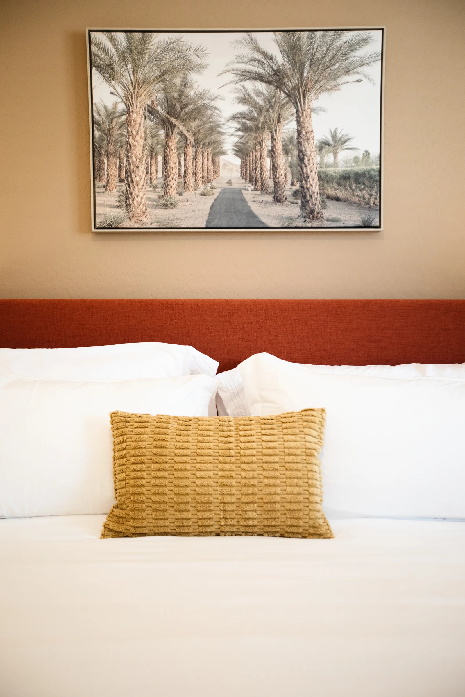 Minimalist bedroom with a rust-colored headboard, a golden textured pillow, and framed art of a palm tree-lined pathway.