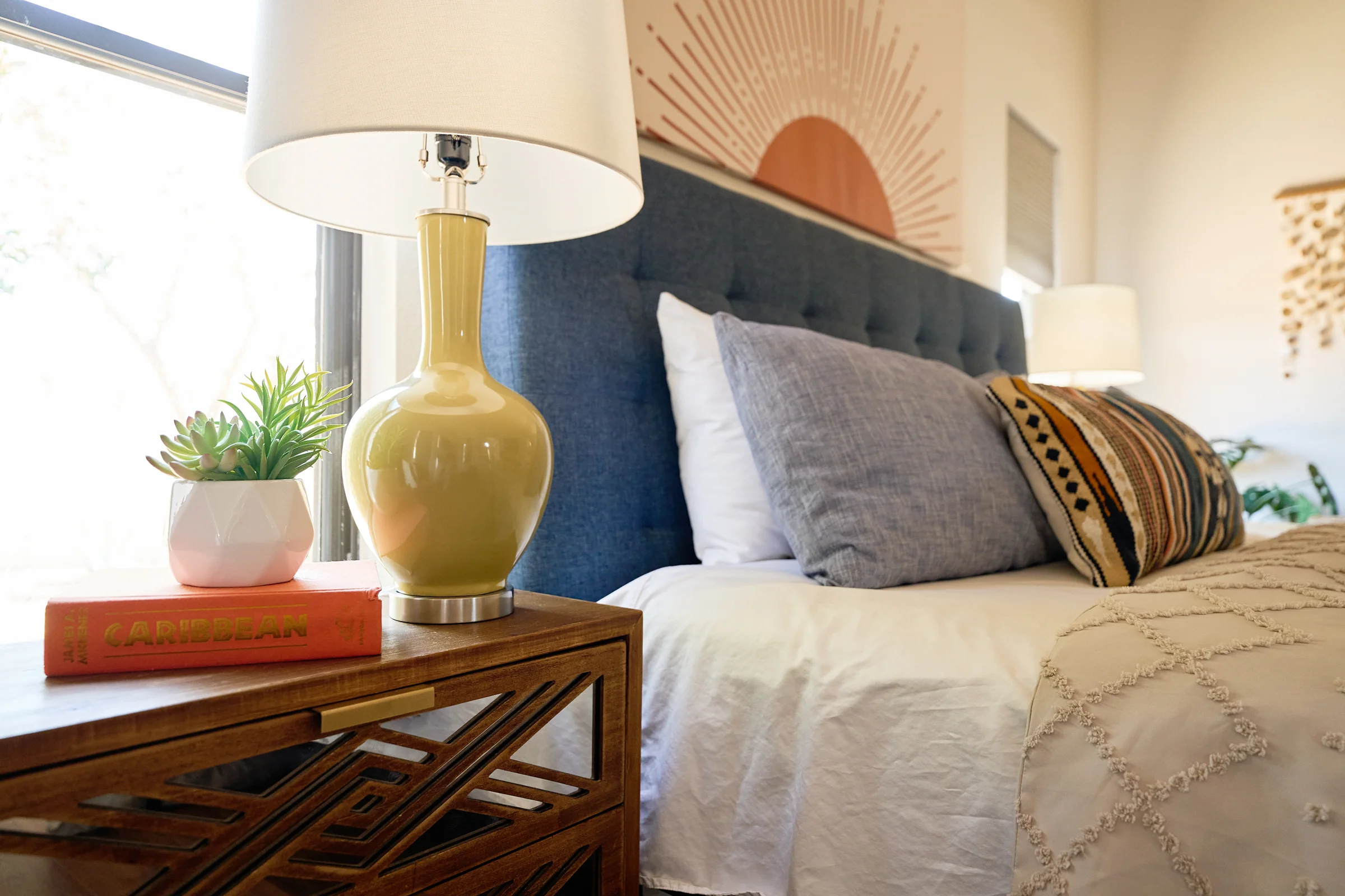 Close-up of a styled bedside table with a geometric lamp, succulent plant, and decorative book, next to a bed with a blue tufted headboard.