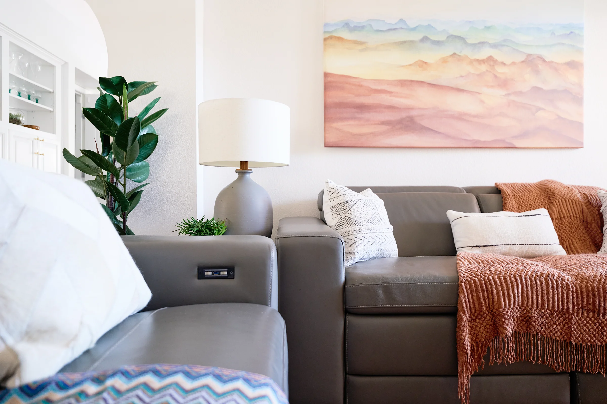 Modern living room with a gray leather sofa, neutral pillows, a ceramic lamp, and a colorful abstract mountain painting on the wall.