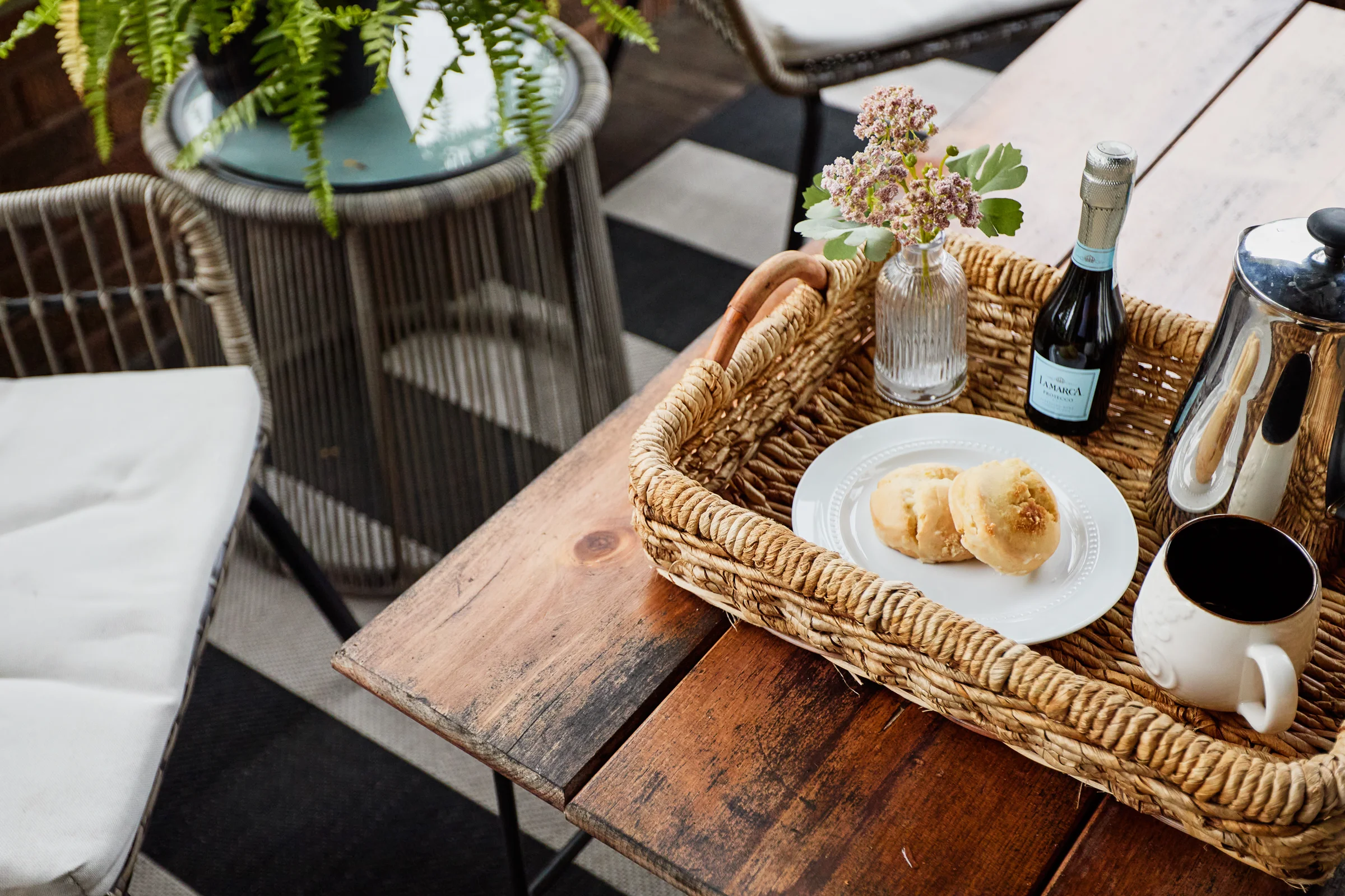 A rustic breakfast tray with pastries, coffee, and a small bottle of sparkling wine, placed on a wooden table in a cozy outdoor setting.