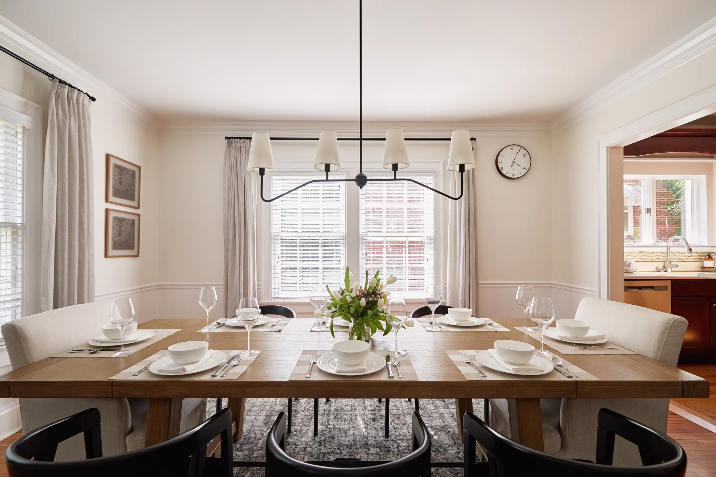 A bright and elegant dining room with a wooden table set for six, featuring white dinnerware, modern black chairs, and a central floral arrangement. Captured by Shane Baker Studios.