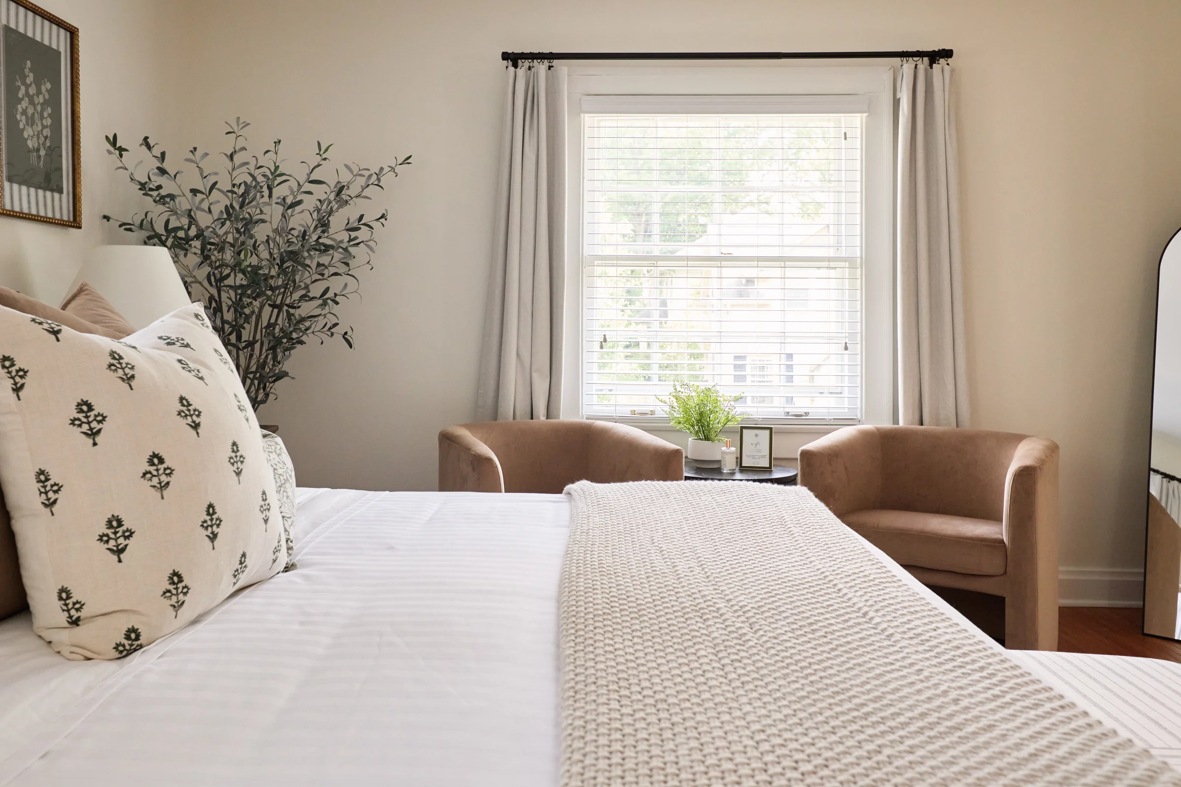 Bright and inviting bedroom with neutral tones, featuring a cozy bed, soft beige chairs, and a large window with white curtains.