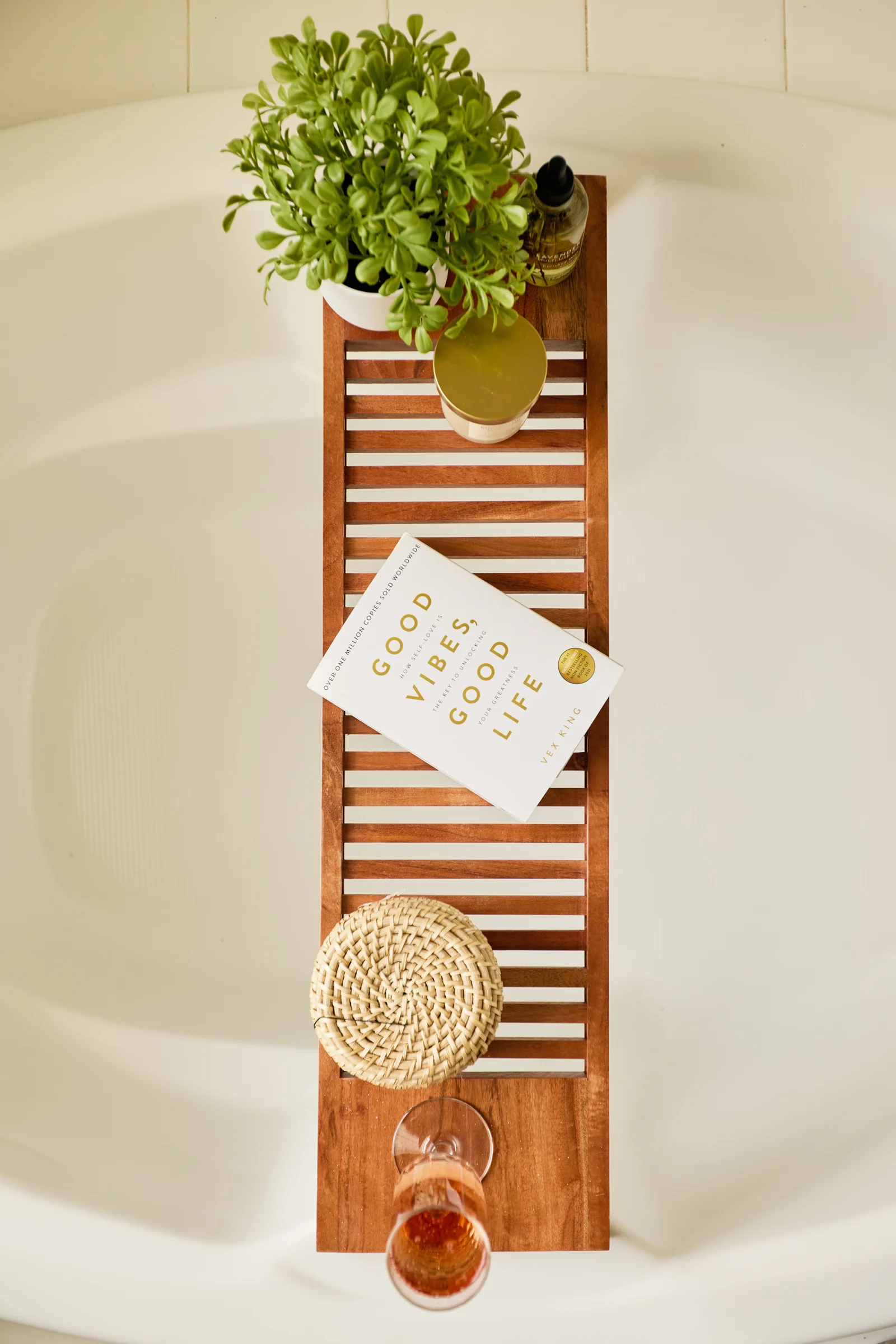 A relaxing bathtub scene with a wooden bath tray holding a plant, a candle, a book titled "Good Vibes, Good Life," and a glass of rosé.