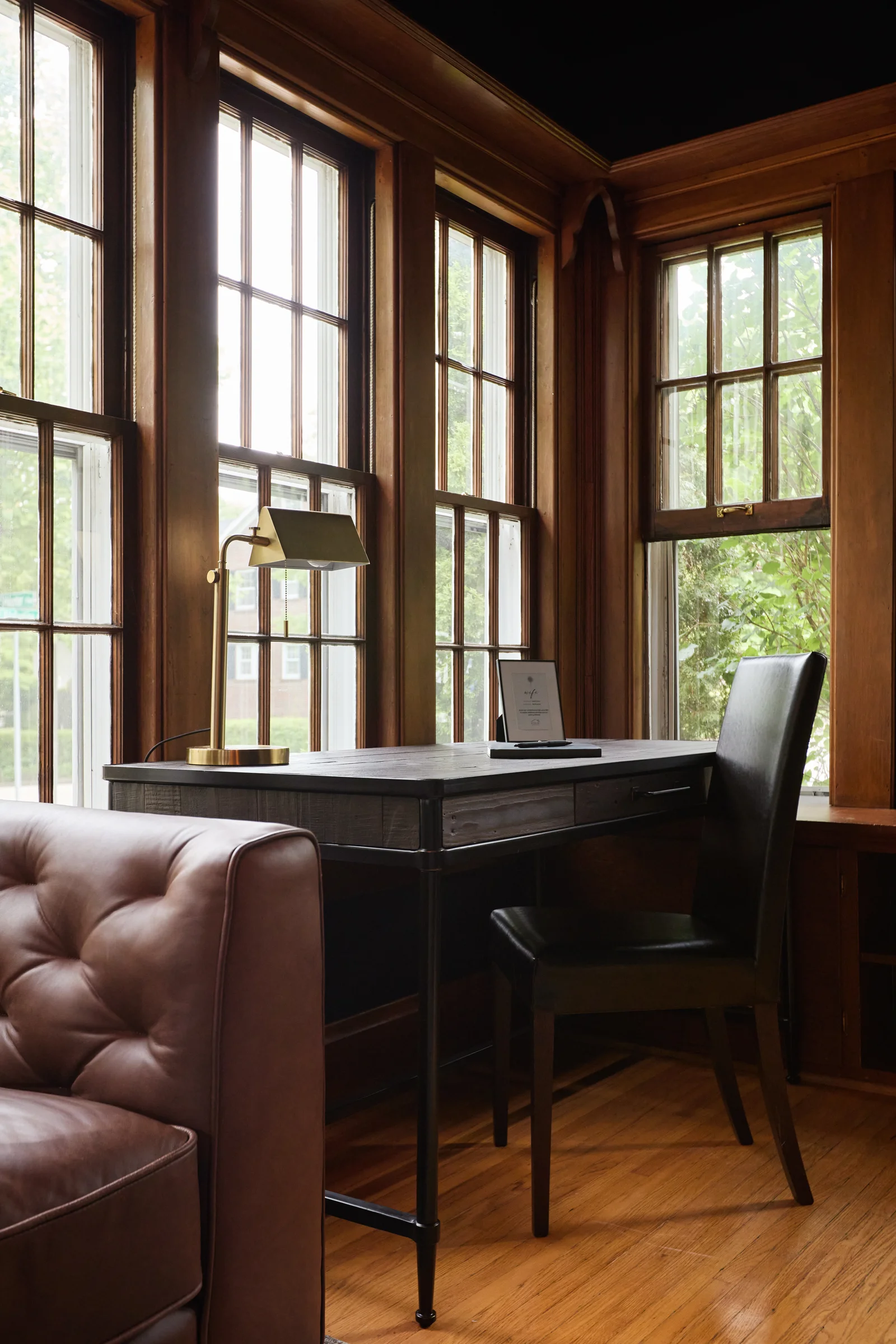 A cozy workspace featuring a dark wooden desk, a sleek black chair, and a brass desk lamp, surrounded by large windows with natural light. Photographed by Shane Baker Studios.