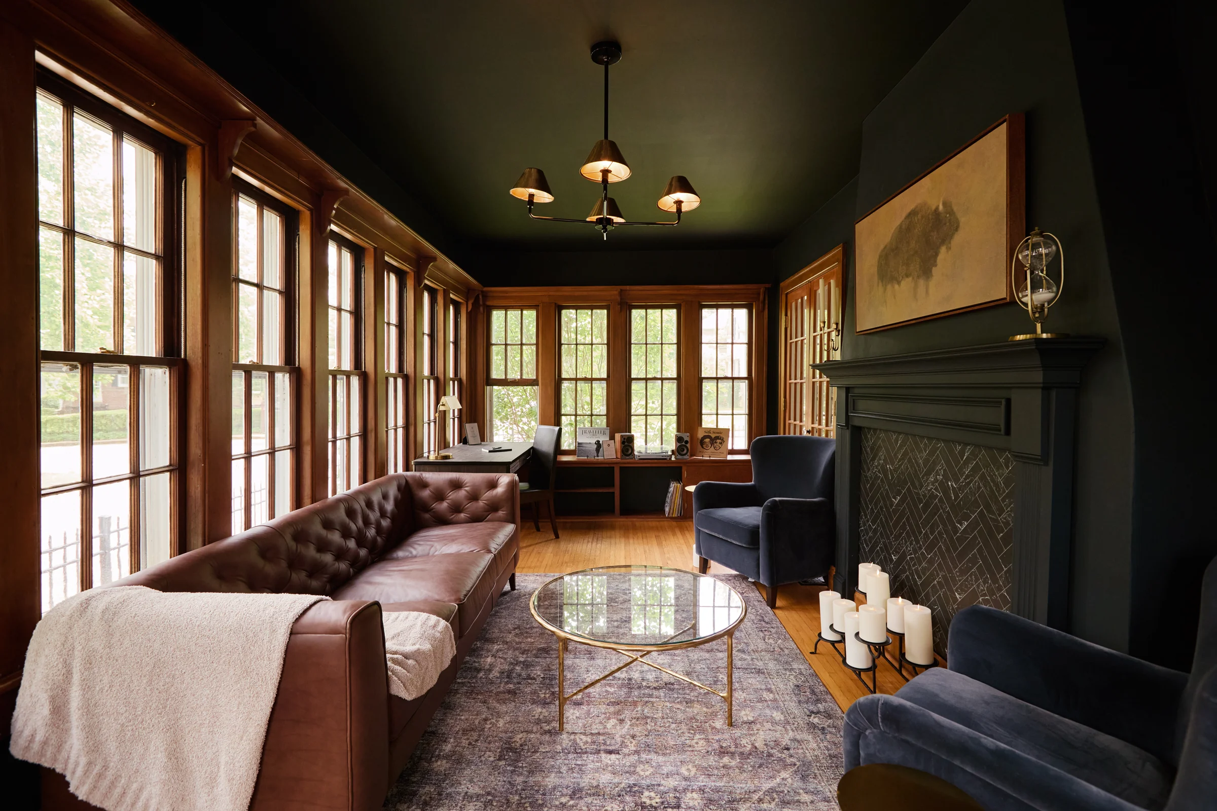 A sophisticated sitting room with dark green walls, a tufted leather sofa, blue velvet chairs, and a fireplace accented by a herringbone tile pattern. Large wooden windows provide ample natural light.