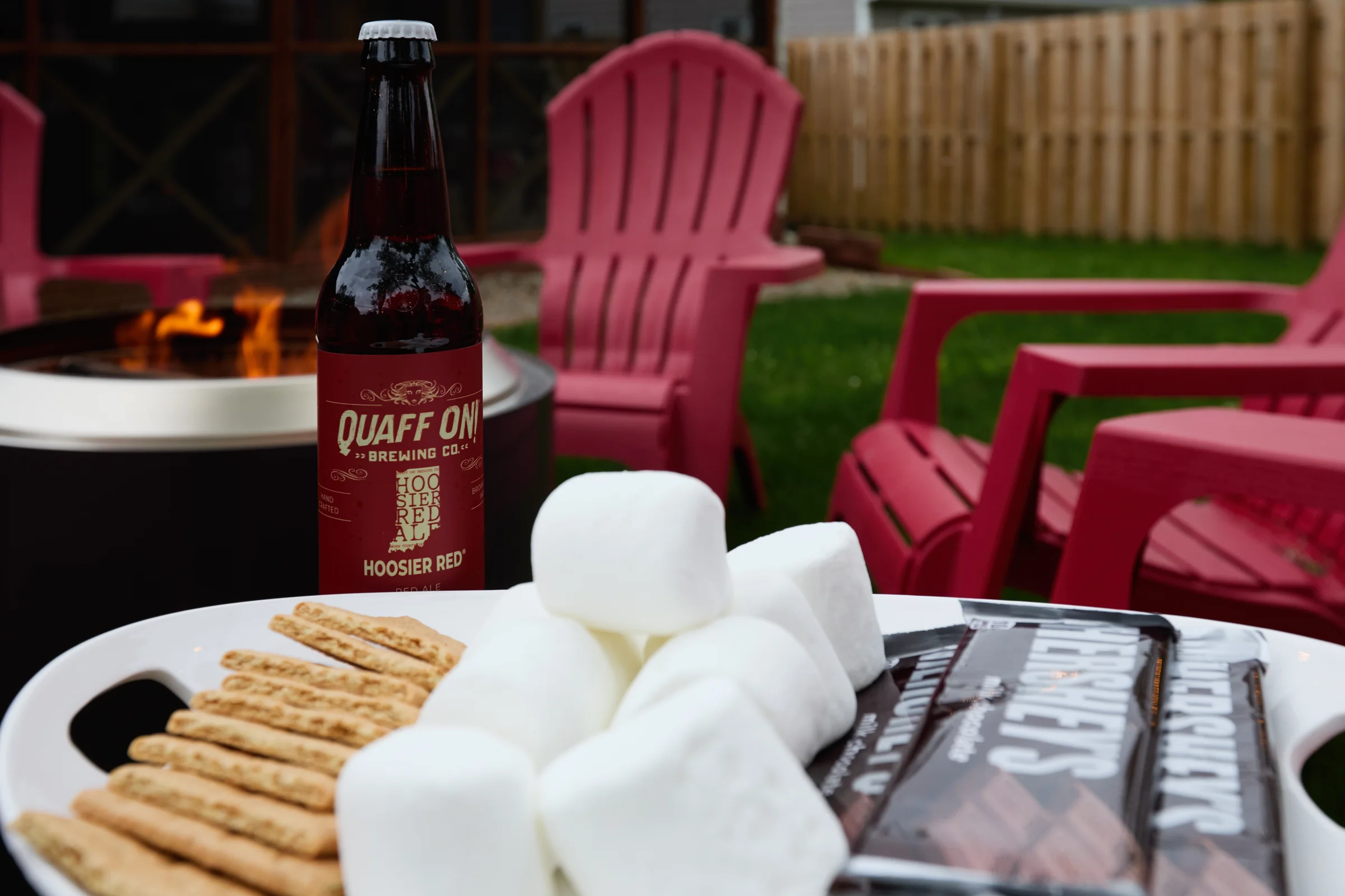 A backyard fire pit setup with marshmallows, chocolate, graham crackers, and a bottle of Quaff On! Brewing Co. Hoosier Red Ale surrounded by red Adirondack chairs. Photographed by Shane Baker Studios.