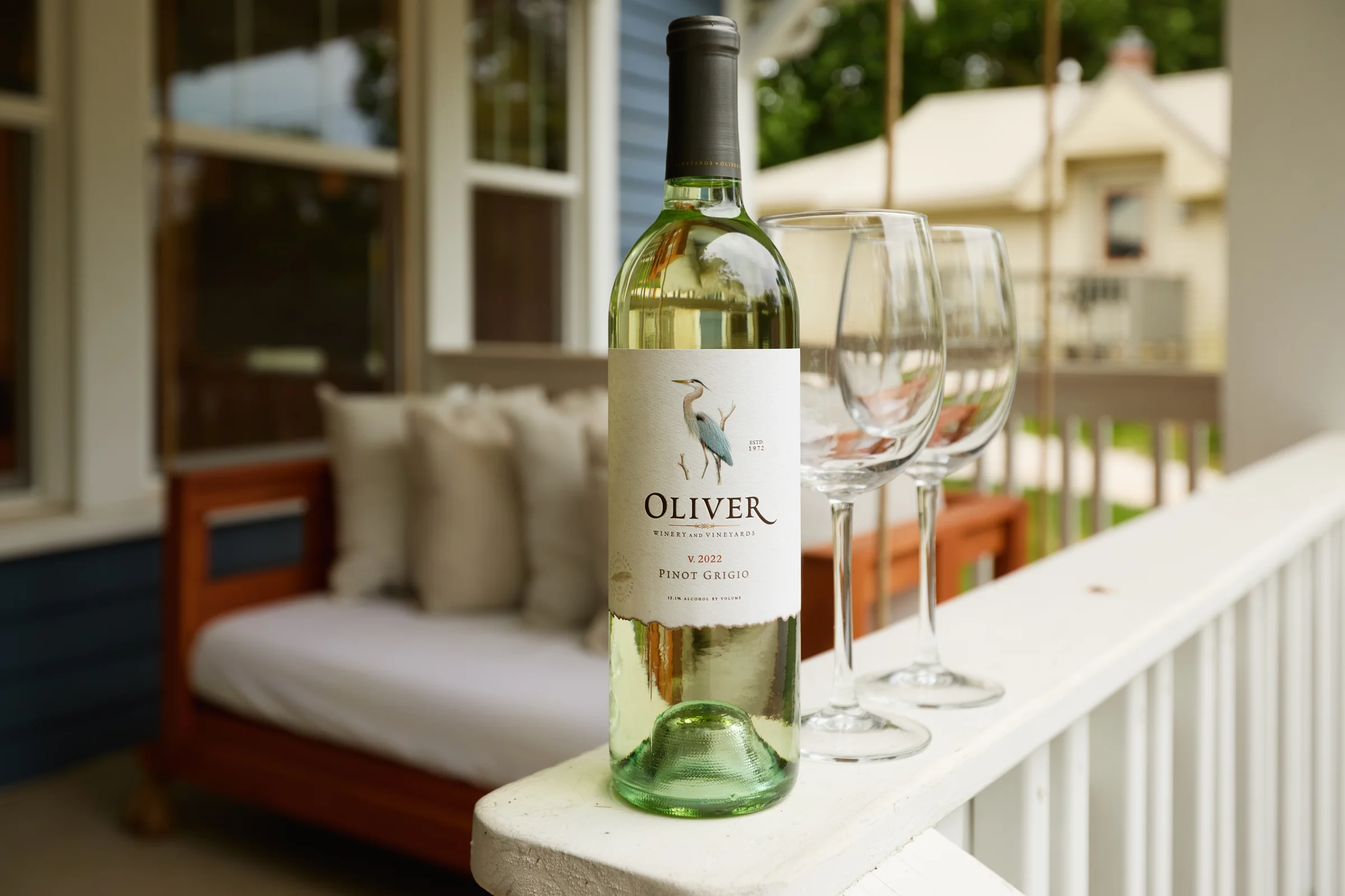 A bottle of Oliver Winery 2022 Pinot Grigio with three wine glasses on a white porch railing, with a cozy swing in the background. Photographed by Shane Baker Studios.