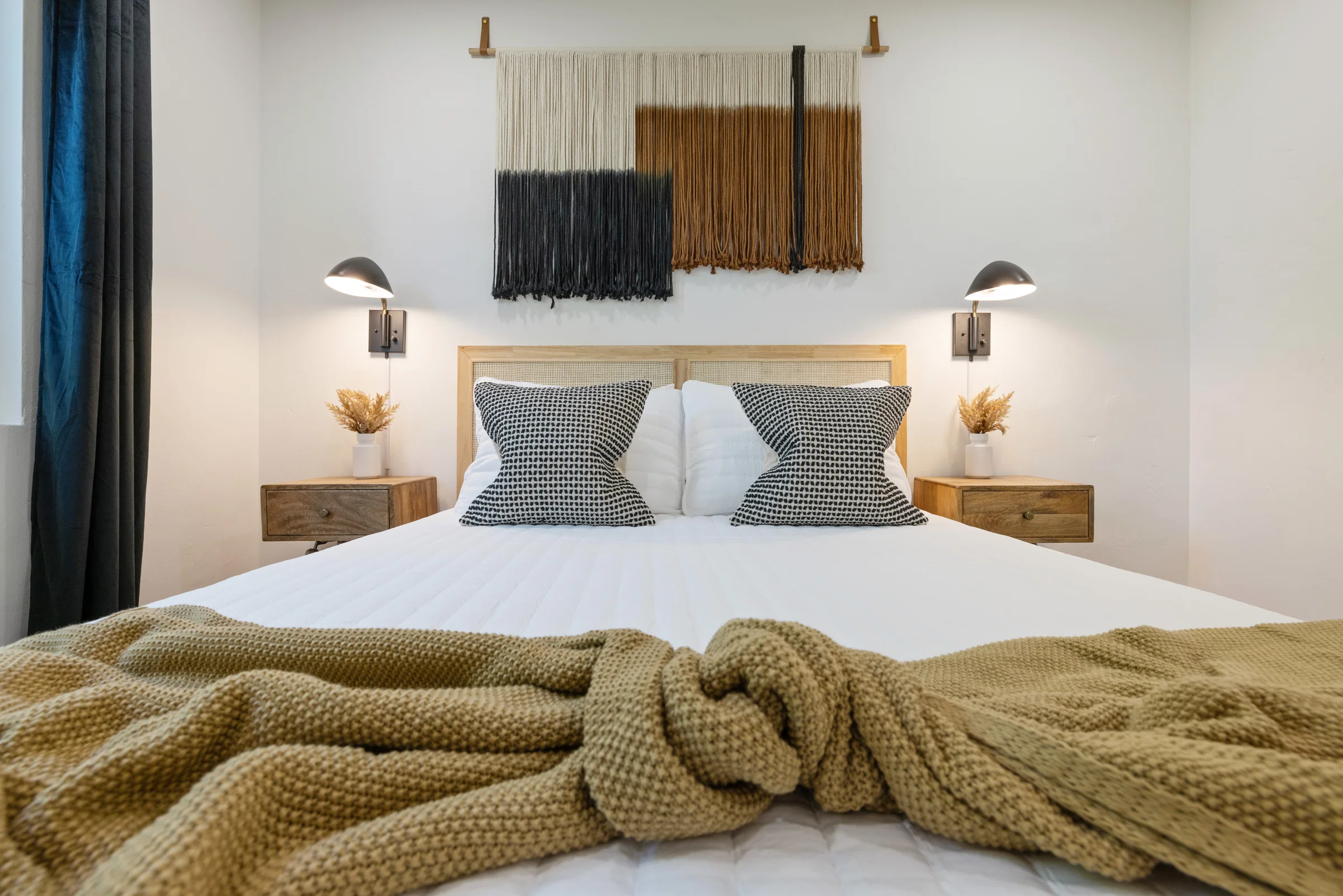 A modern bedroom featuring a cozy bed with black and white patterned pillows, a mustard knit throw blanket, wooden nightstands, and a woven wall hanging above the headboard. Photographed by Shane Baker Studios.