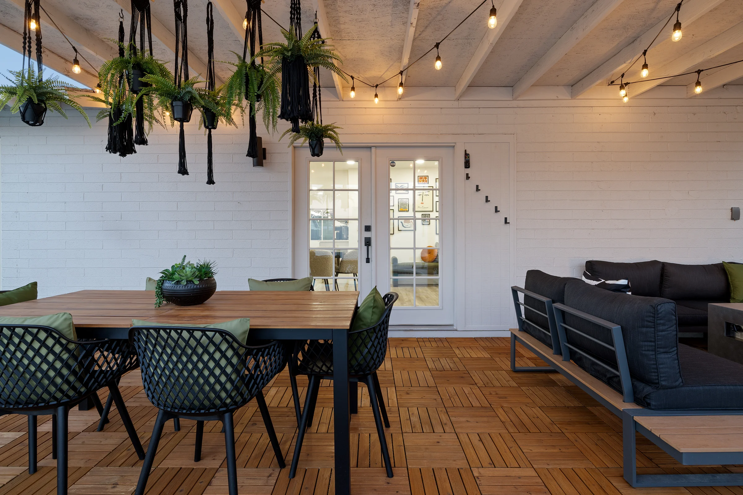 Modern outdoor patio with a wooden dining table, green-accented chairs, black hanging planters with ferns, and string lights under a covered ceiling. Photographed by Shane Baker Studios.