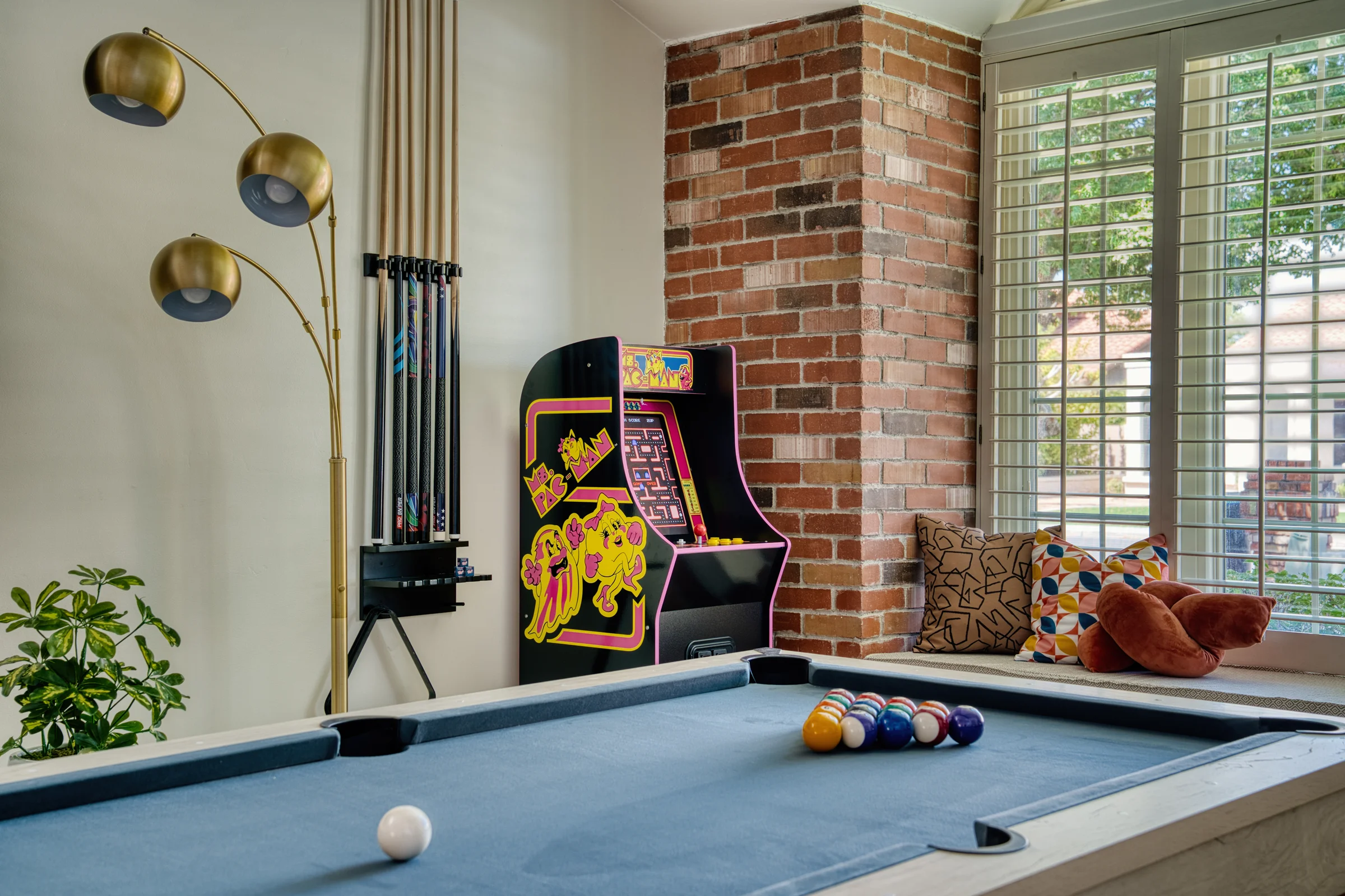 Game room with a blue pool table, a vintage Ms. Pac-Man arcade machine, a gold floor lamp, and brick accents. Photographed by Shane Baker Studios.