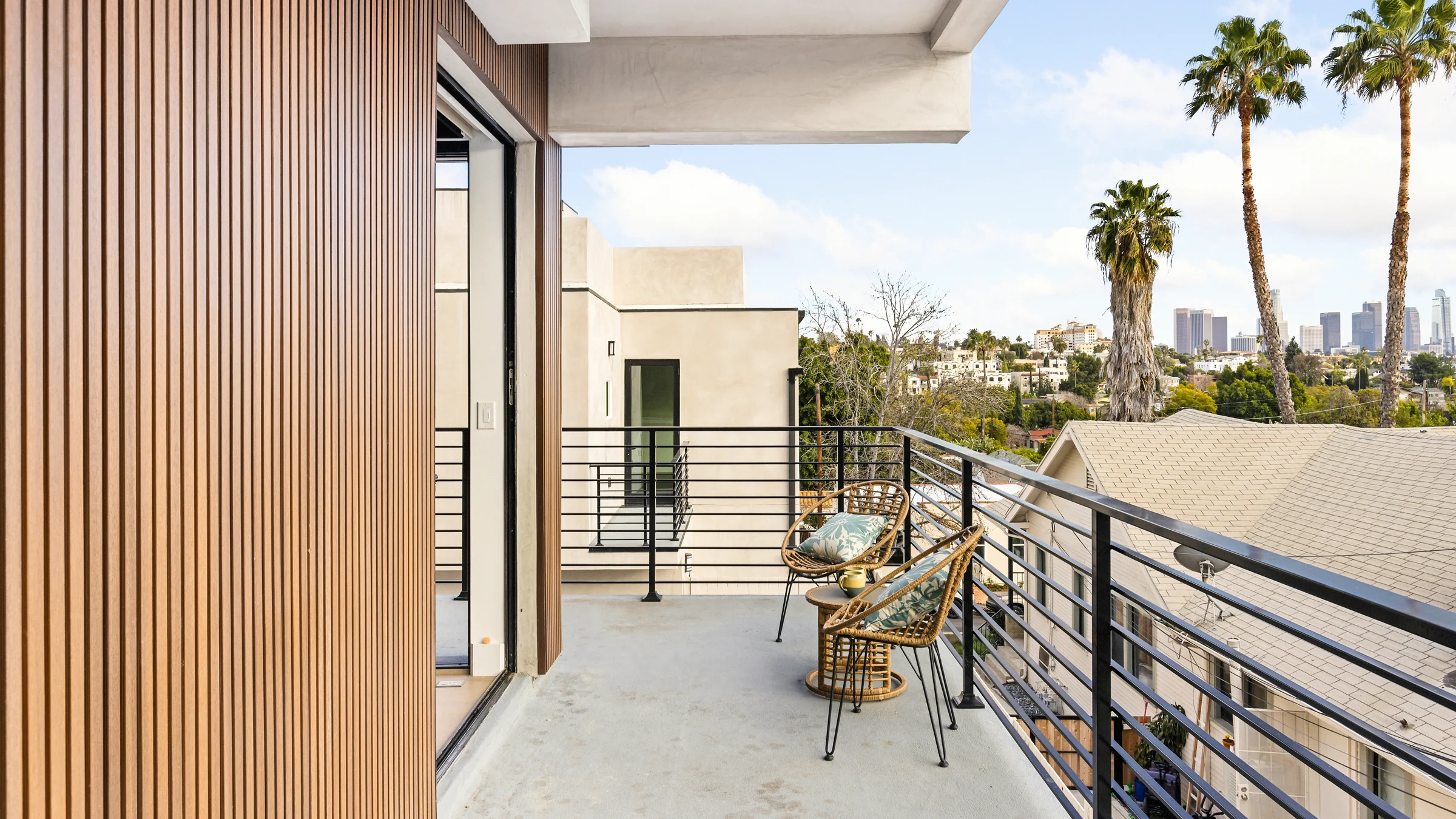 Modern balcony with wood panel accents, wicker chairs, and a view of palm trees and a city skyline. Photographed by Shane Baker Studios.