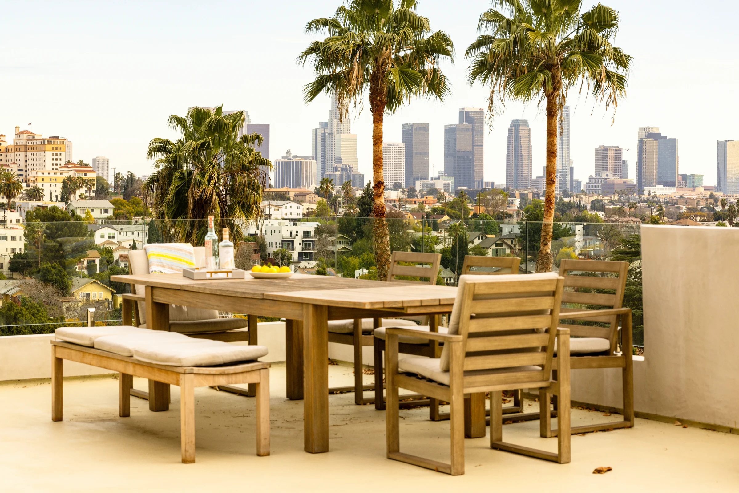 Outdoor rooftop dining area with wooden furniture, palm trees, and a city skyline view in the background.