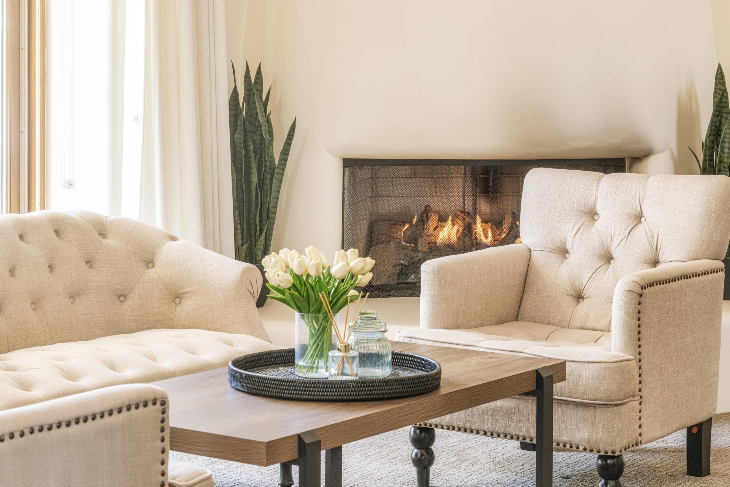 Cozy living room with beige tufted seating, a wooden coffee table adorned with tulips, and a lit fireplace in the background.