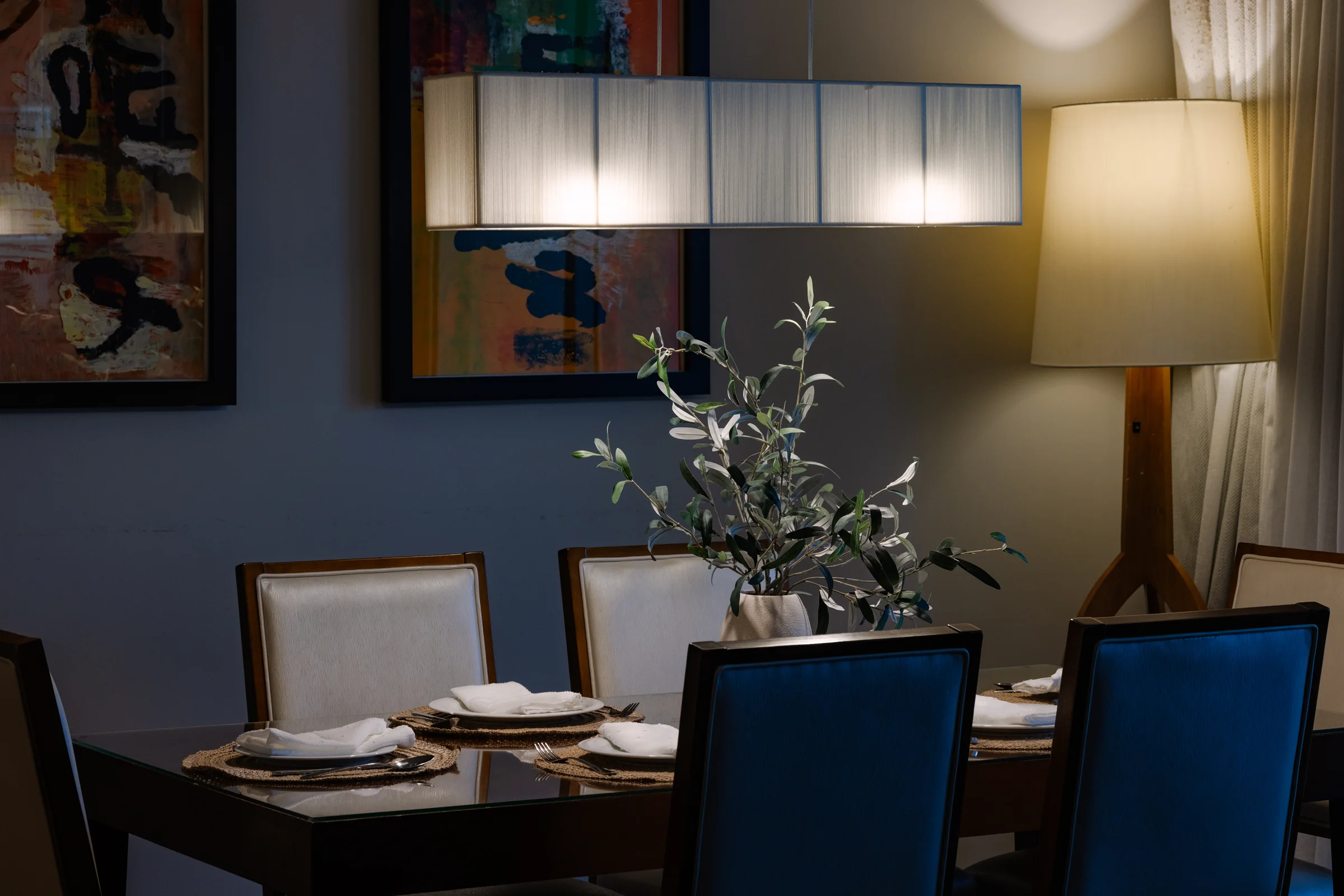 A dining table set with elegant tableware and a centerpiece featuring a vase with olive branches, softly illuminated by modern lighting and surrounded by abstract artwork. Photographed by Shane Baker Studios.