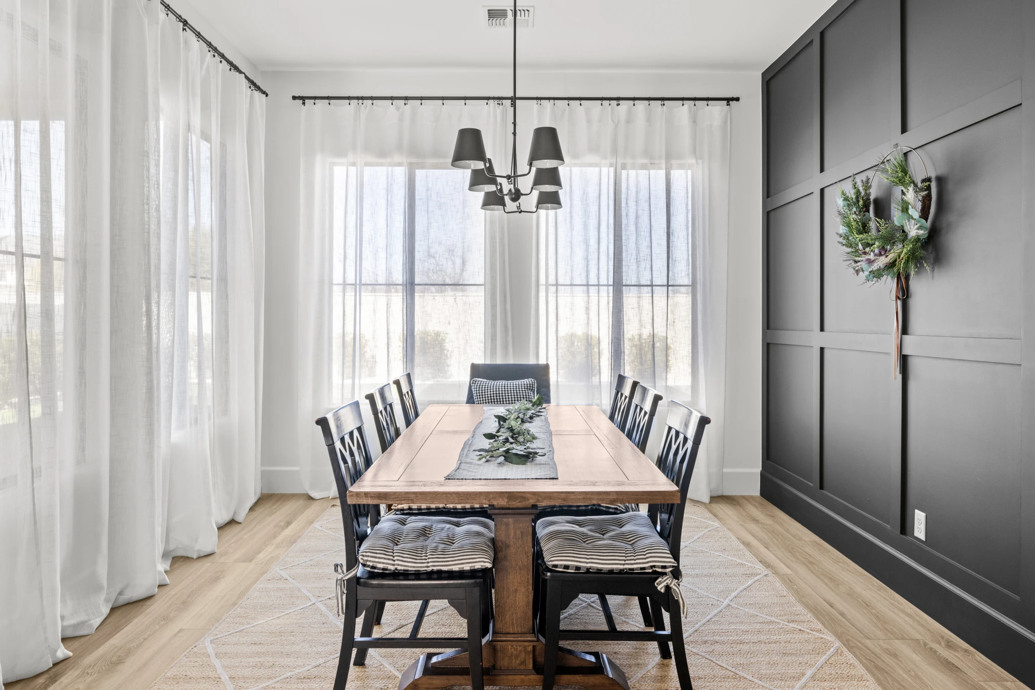 A modern dining room with a wooden table, black chairs, sheer white curtains, and a dark accent wall featuring a wreath. Photographed by Shane Baker Studios.