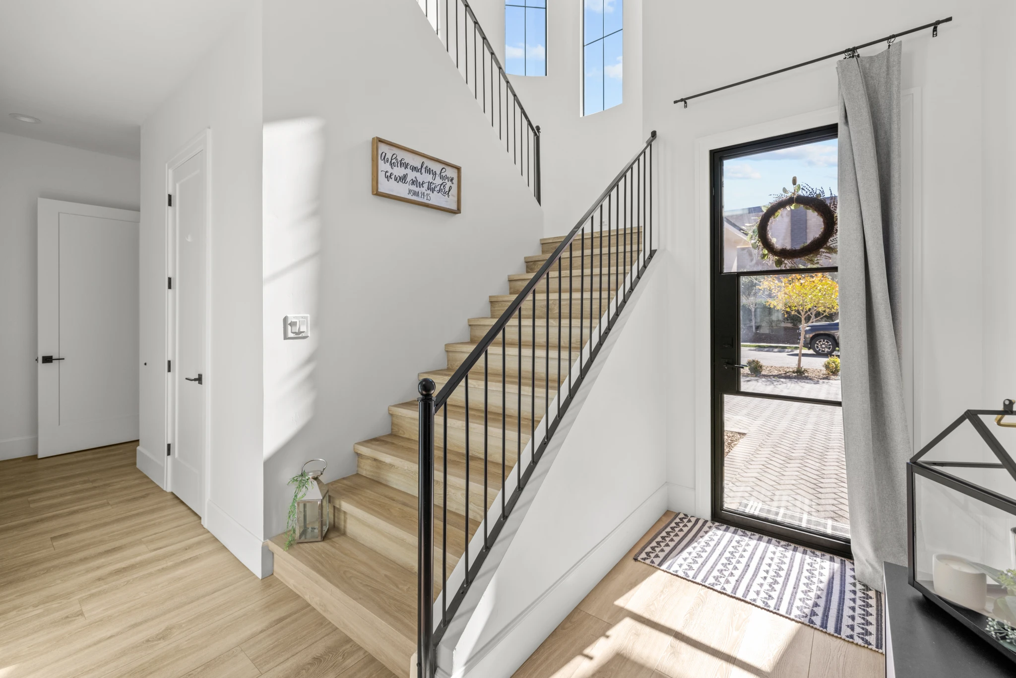 A bright and modern entryway featuring a wooden staircase with black metal railing, a glass-paneled front door, and natural light streaming in. Photographed by Shane Baker Studios.