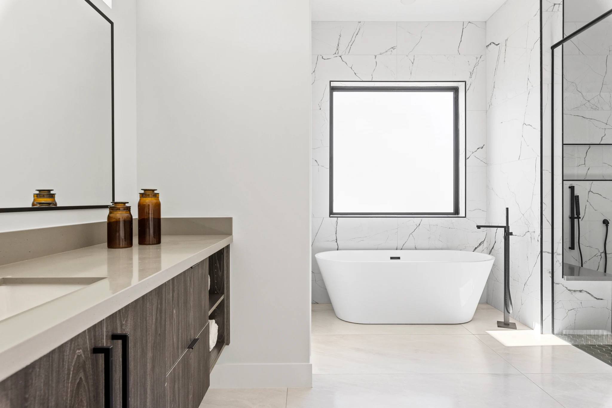 A modern primary bathroom featuring a floating vanity, a freestanding soaking tub, and a glass-enclosed shower with marble-inspired tile. Photographed by Shane Baker Studios.