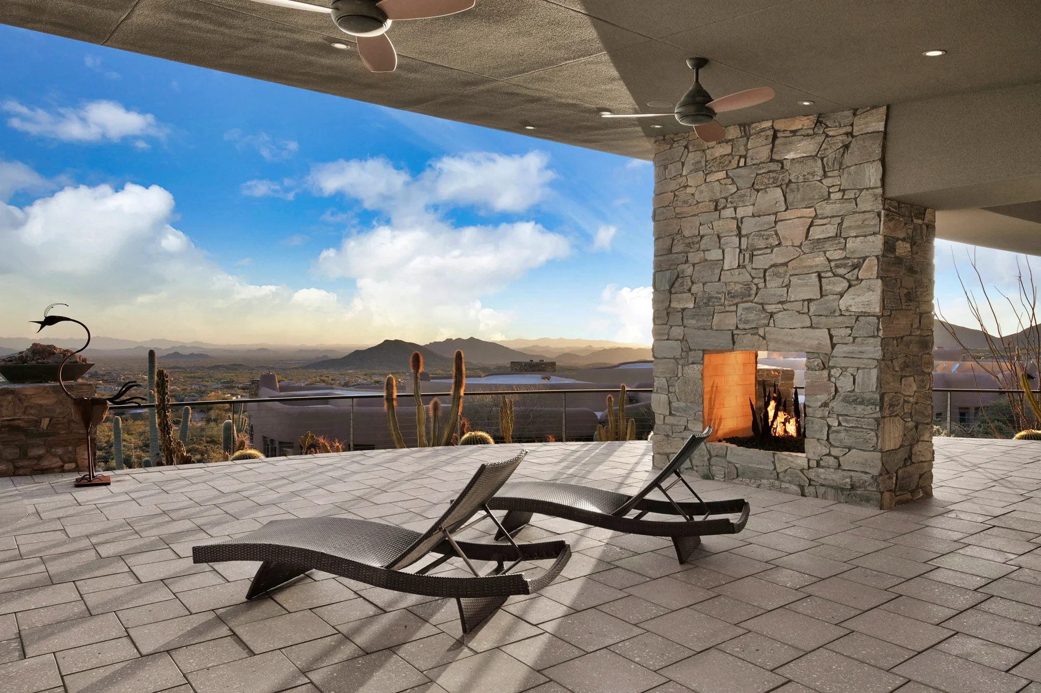 A luxurious outdoor patio with a stone fireplace, two lounge chairs, desert landscaping, and a panoramic view of mountains under a blue sky.