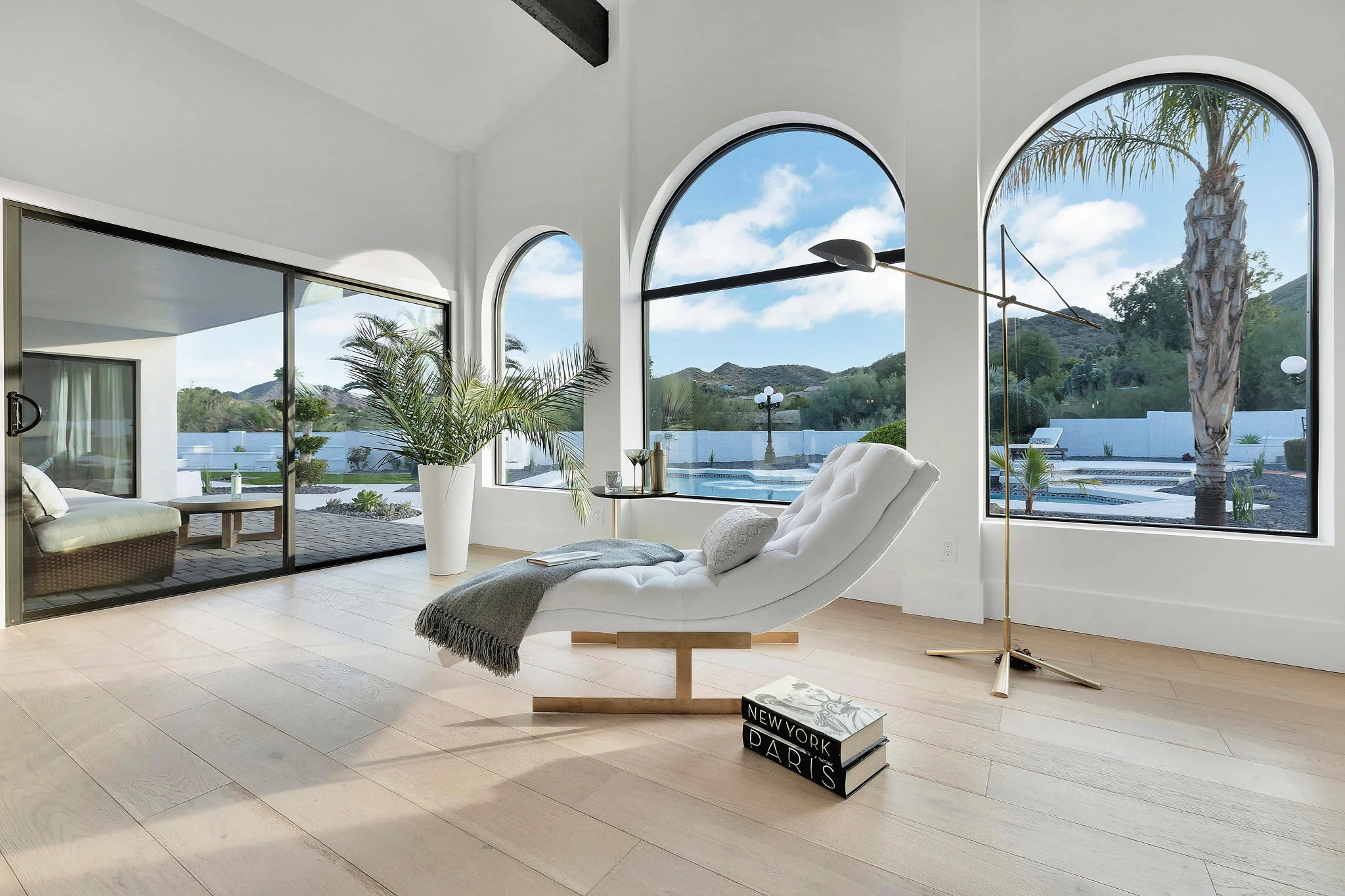 Modern living space with large arched windows, a white chaise lounge, and a view of palm trees and hills outside.