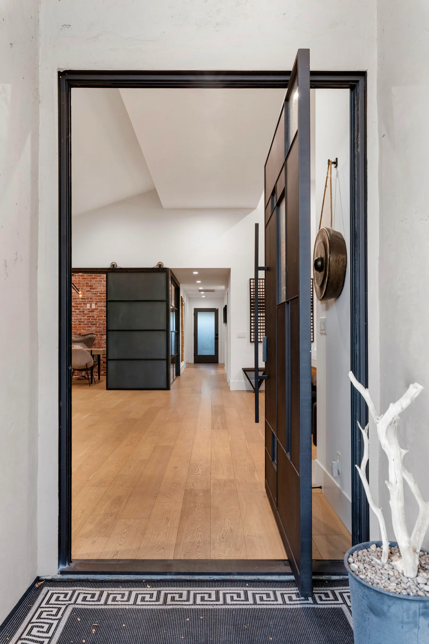 A modern entryway featuring a metal-framed pivot door, exposed brick accents, and warm wood flooring.