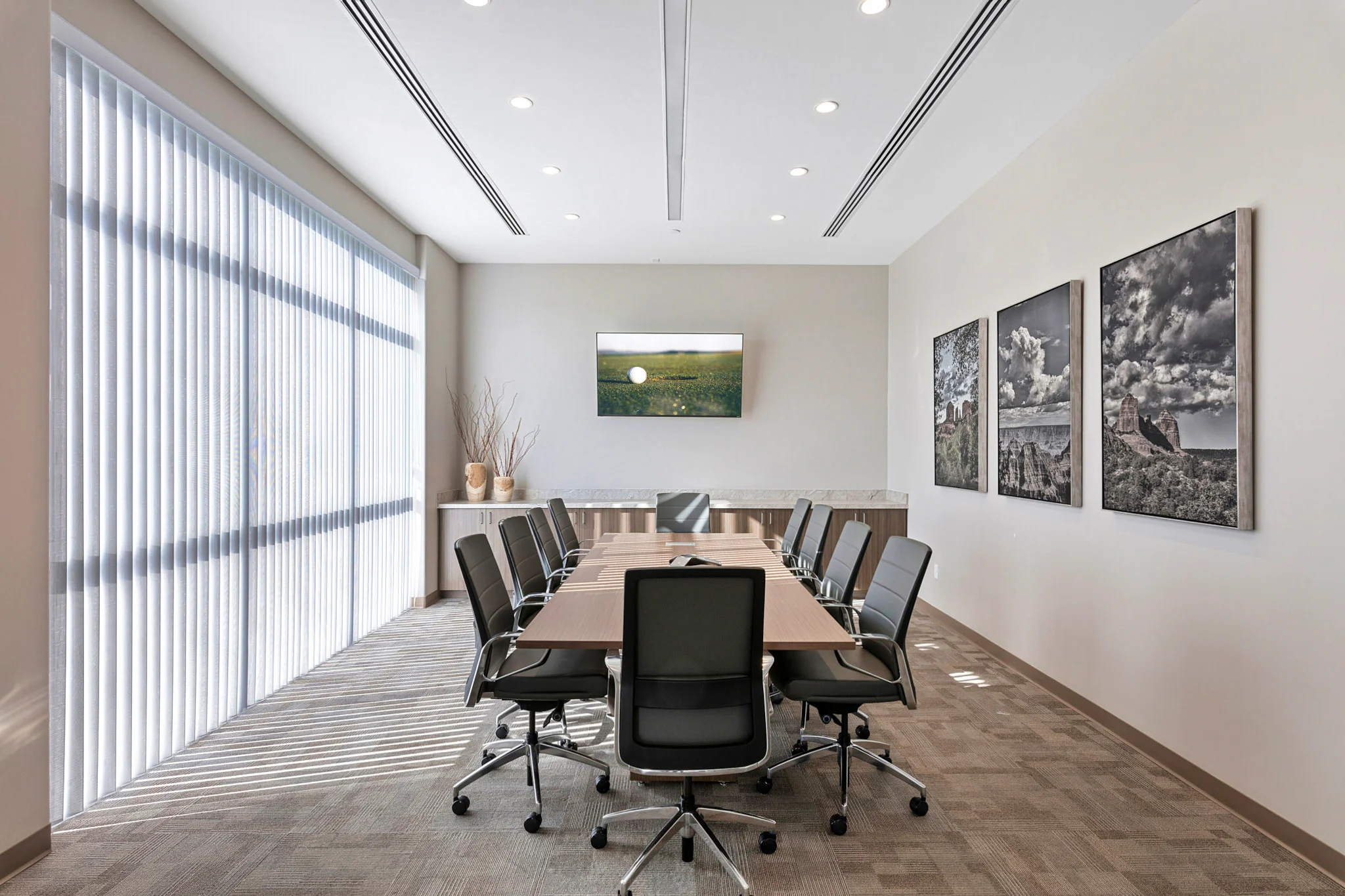 A modern conference room featuring a long wooden table with black swivel chairs, floor-to-ceiling windows with vertical blinds, and scenic artwork on the walls.