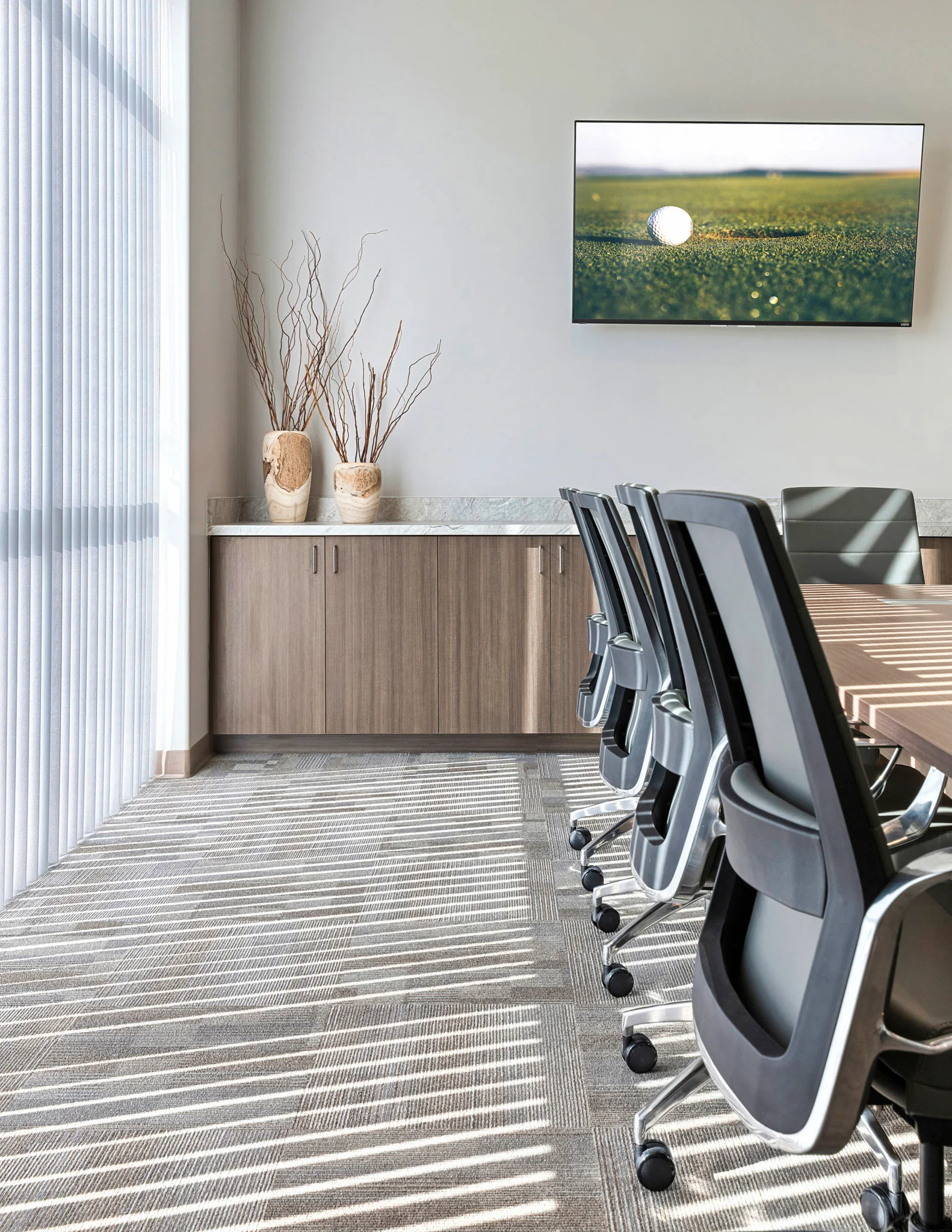 Modern conference room with sleek black chairs, a wooden table, natural light streaming through vertical blinds, and a TV displaying a golf ball on grass.