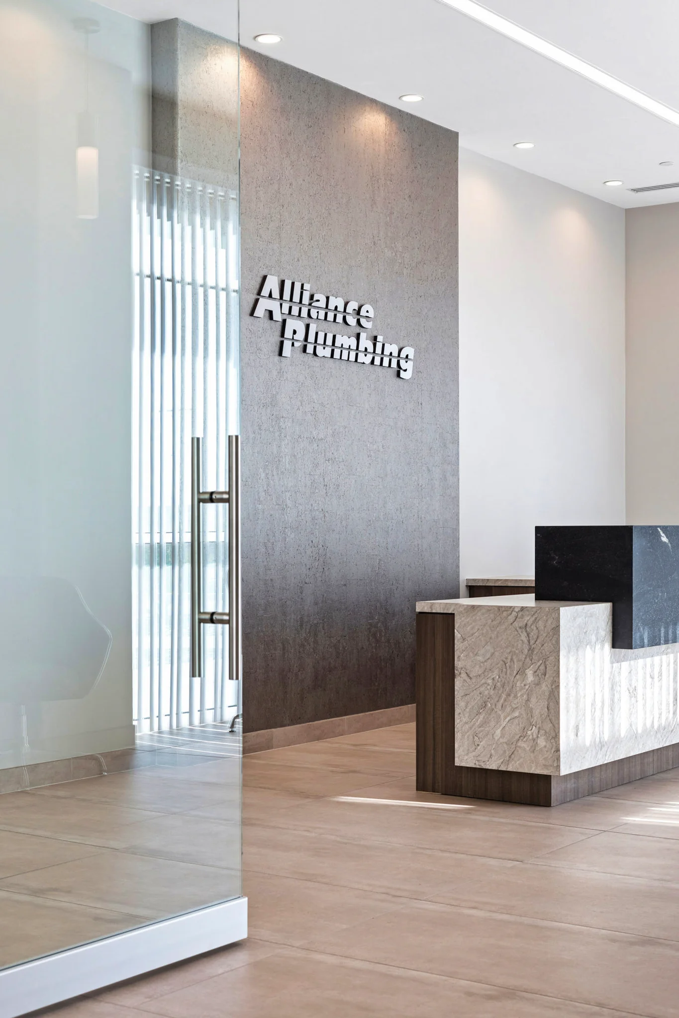 Modern reception area for Alliance Plumbing, featuring a textured accent wall with 3D signage, a glass entry door, and a stone-faced reception desk.