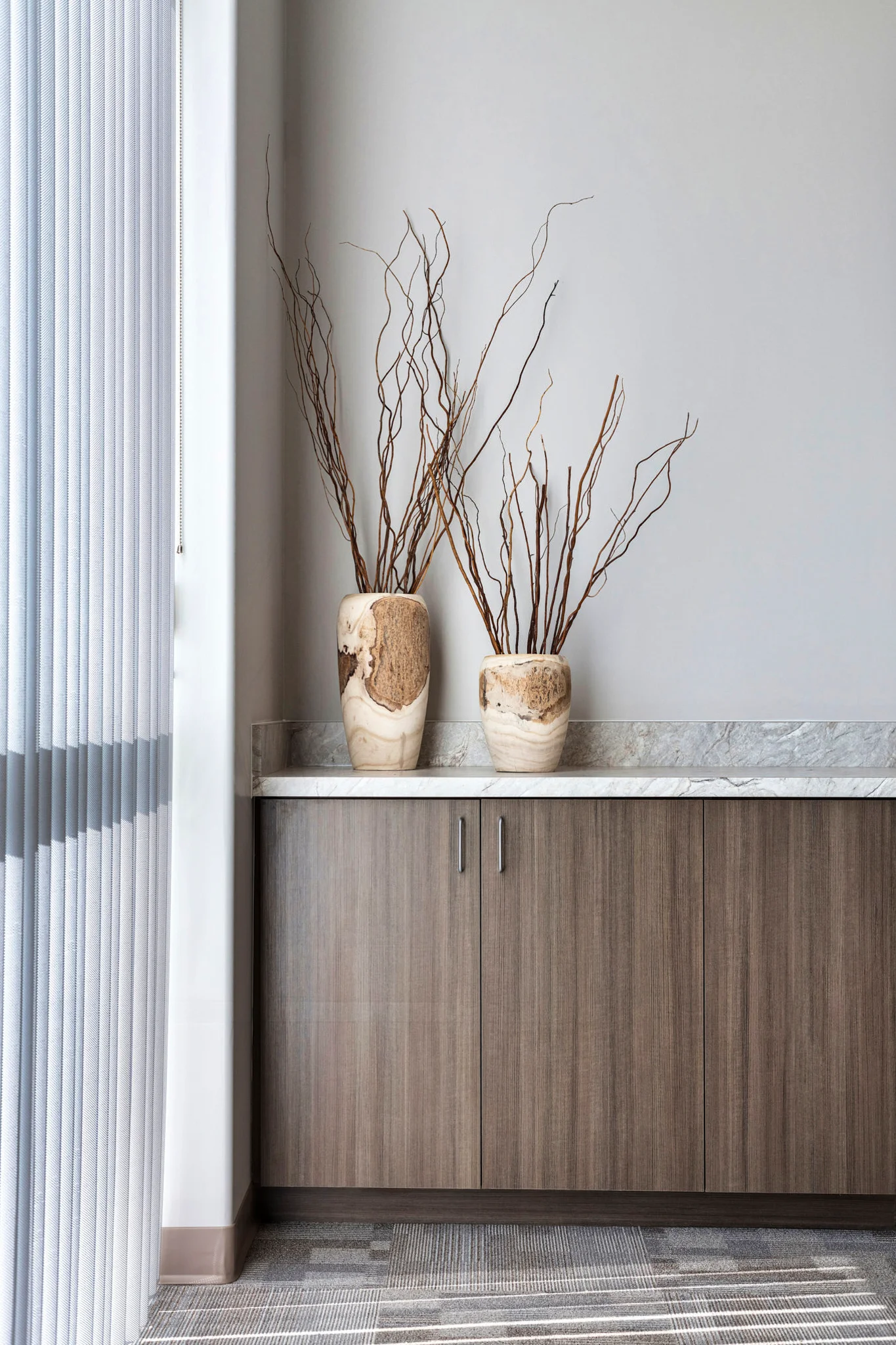 Minimalist decor featuring two stone vases with natural wood branches on a marble countertop atop a wooden cabinet, next to vertical blinds.