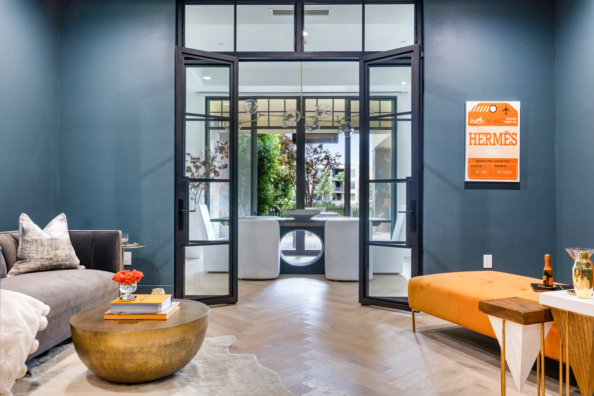 A stylish living room featuring a deep teal accent wall, a modern gold coffee table, and double glass doors leading to a bright dining area, captured by Shane Baker Studios.