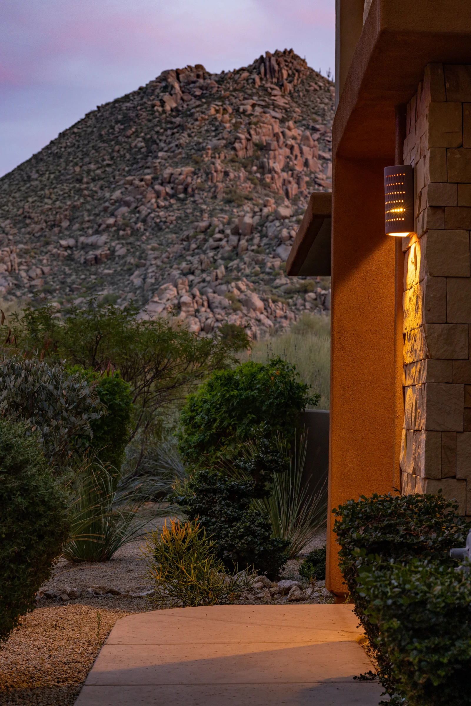 Evening view of a desert landscape with a stone and stucco house illuminated by a modern outdoor light fixture, surrounded by lush desert plants. Photographed by Shane Baker Studios.