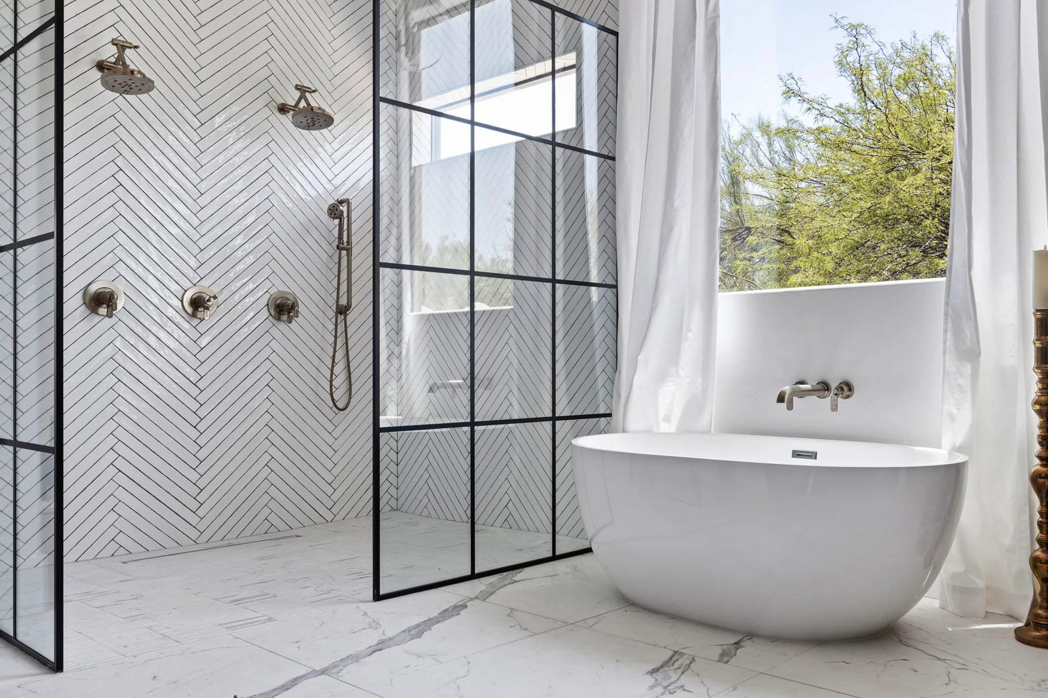 A luxurious bathroom with a herringbone tile shower, black-framed glass partitions, and a freestanding white bathtub near a large window, photographed by Shane Baker Studios.