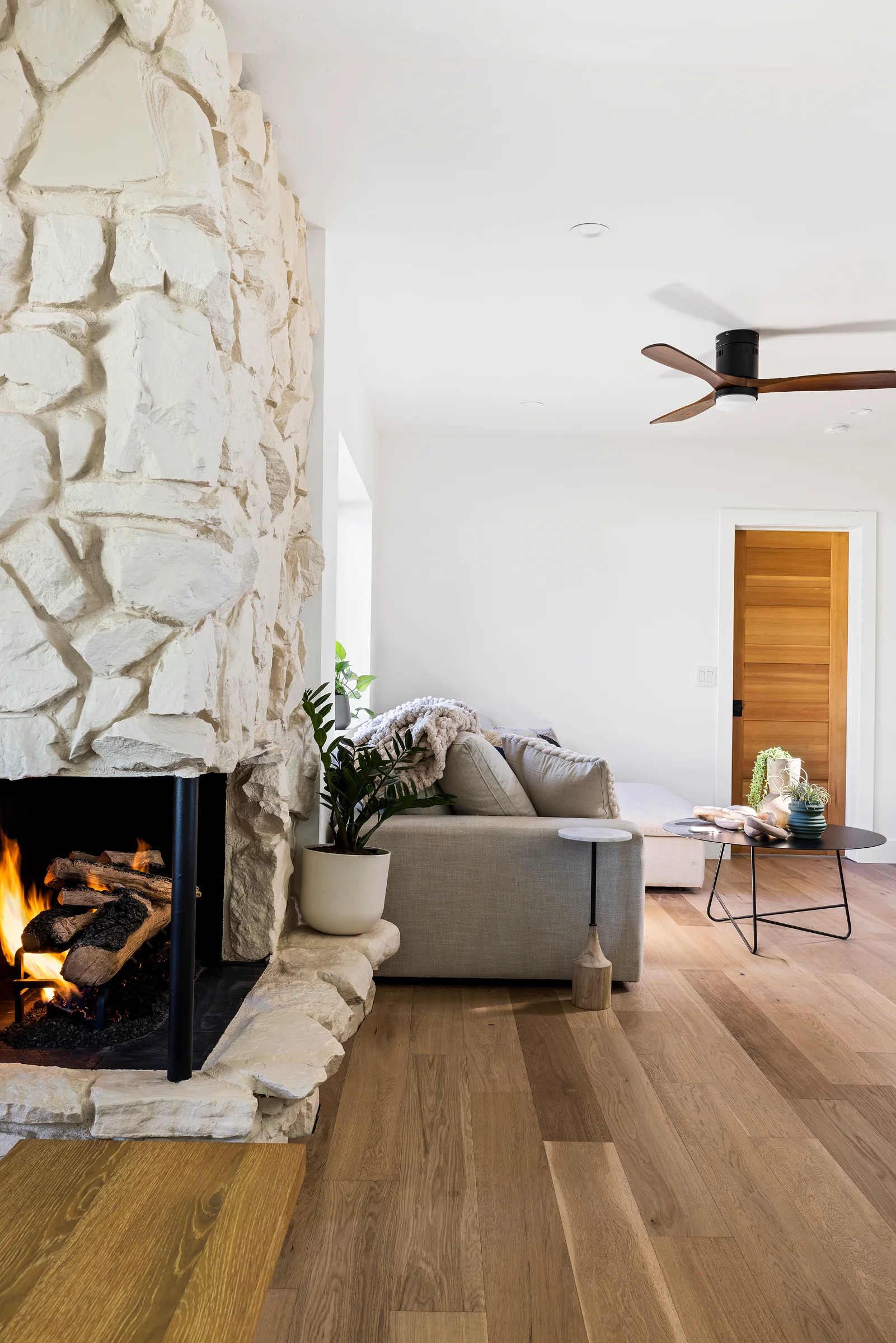 Cozy living room with a stone fireplace, modern ceiling fan, and neutral-toned decor featuring a sofa and indoor plant.