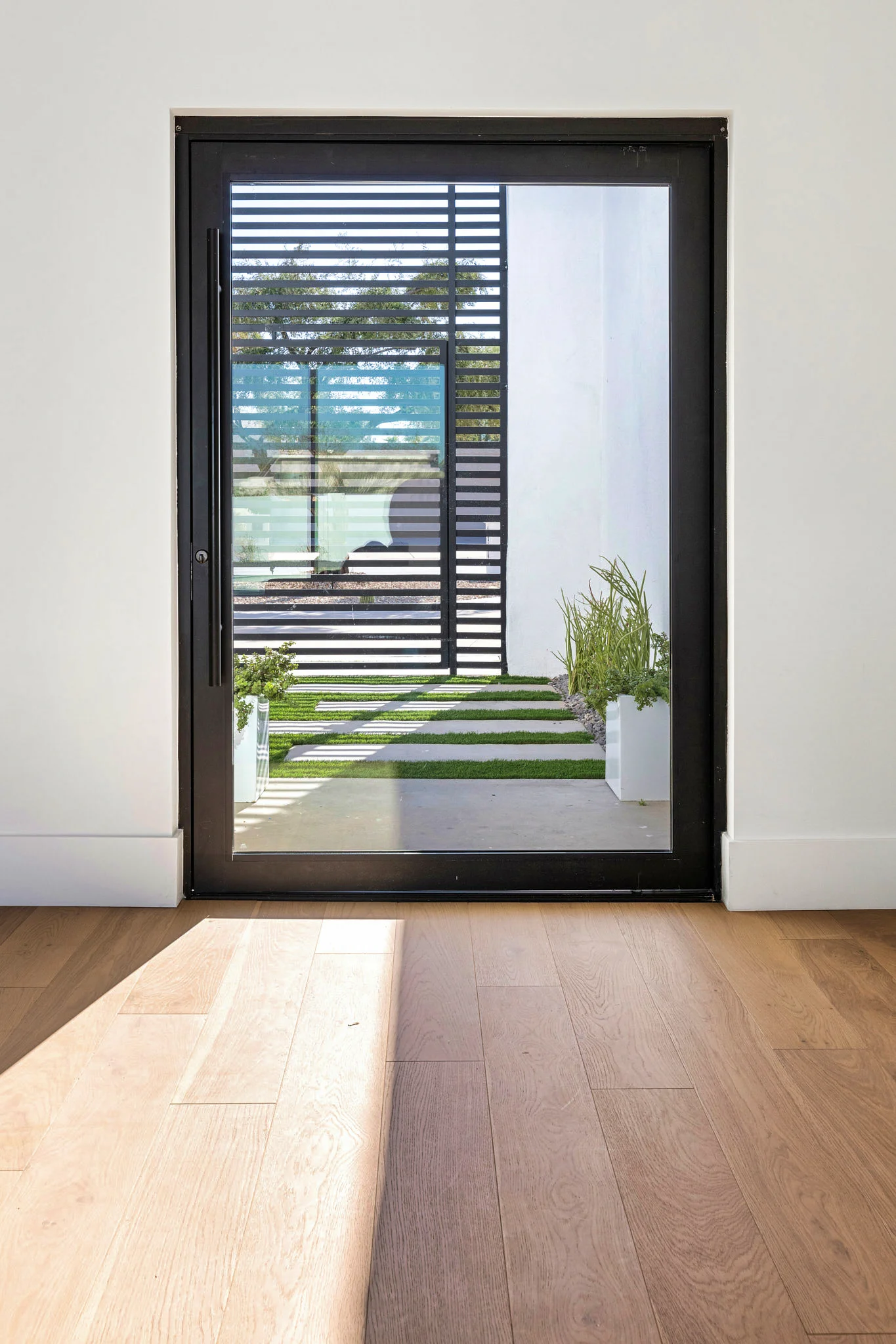 View through a black-framed glass door showcasing a minimalist courtyard with alternating concrete slabs and grass, framed by a modern privacy screen.