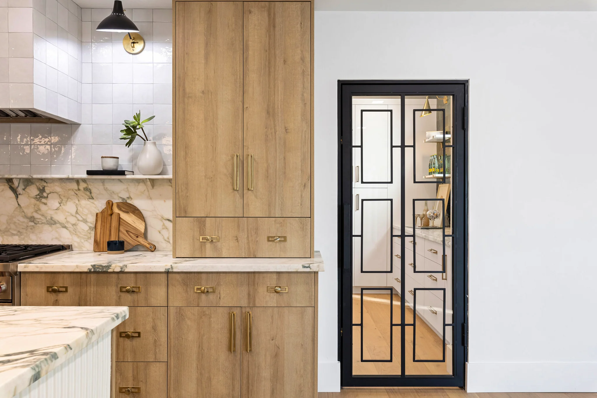 A modern kitchen with natural wood cabinets, marble countertops, and a sleek pantry door with geometric black framing.