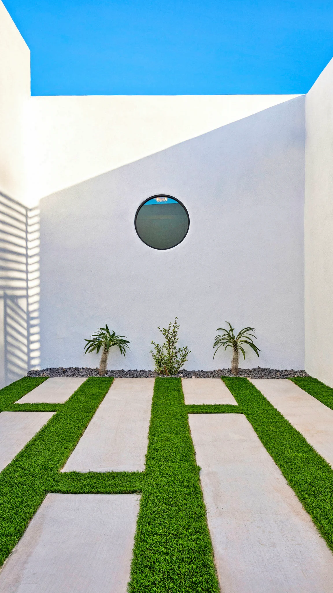 Minimalist courtyard with white walls, a circular window, bright blue sky, and a patterned walkway of concrete slabs and green grass strips.