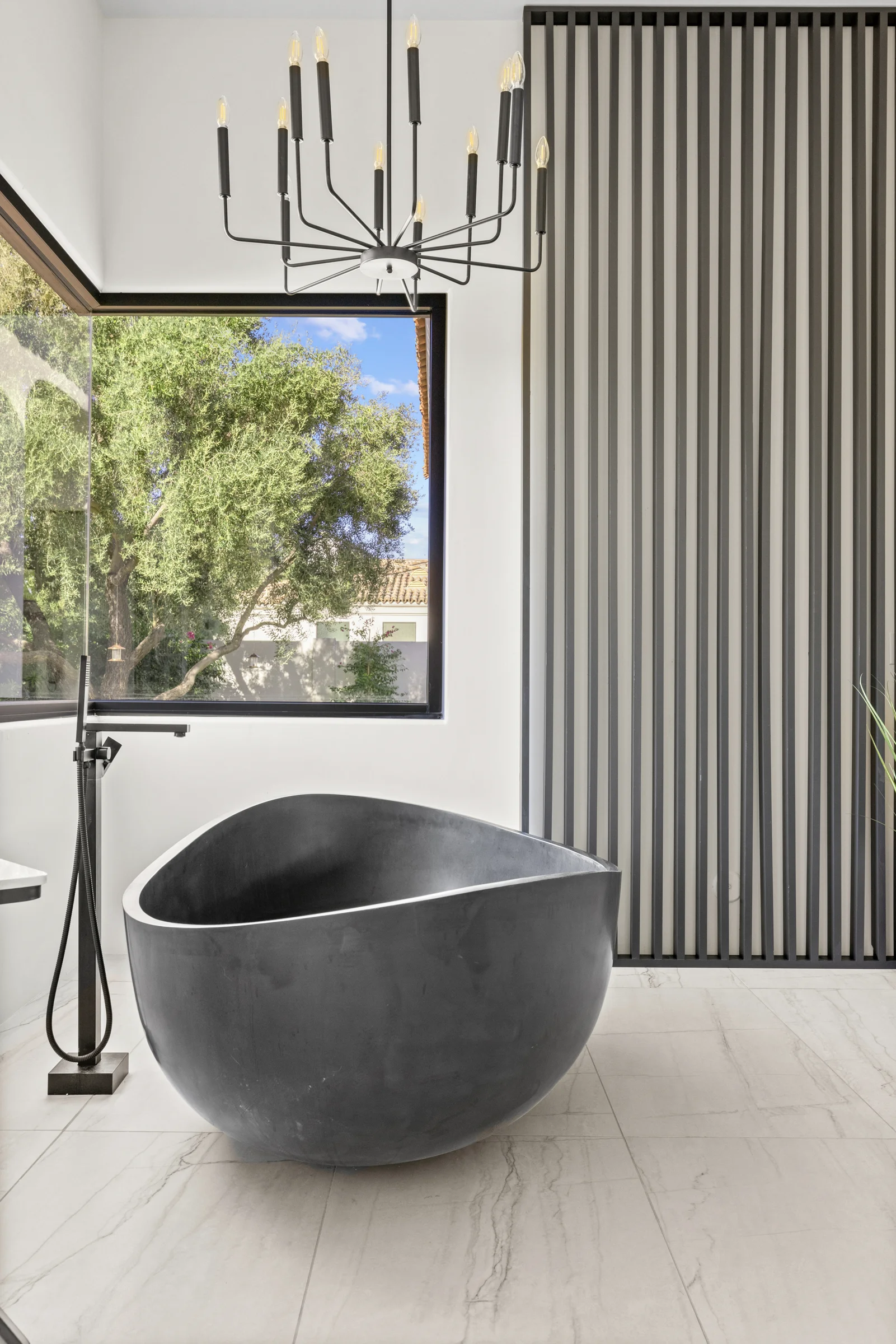 Modern bathroom featuring a sculptural black freestanding bathtub, minimalist black fixtures, a large window with a view of greenery, and a striking black chandelier.