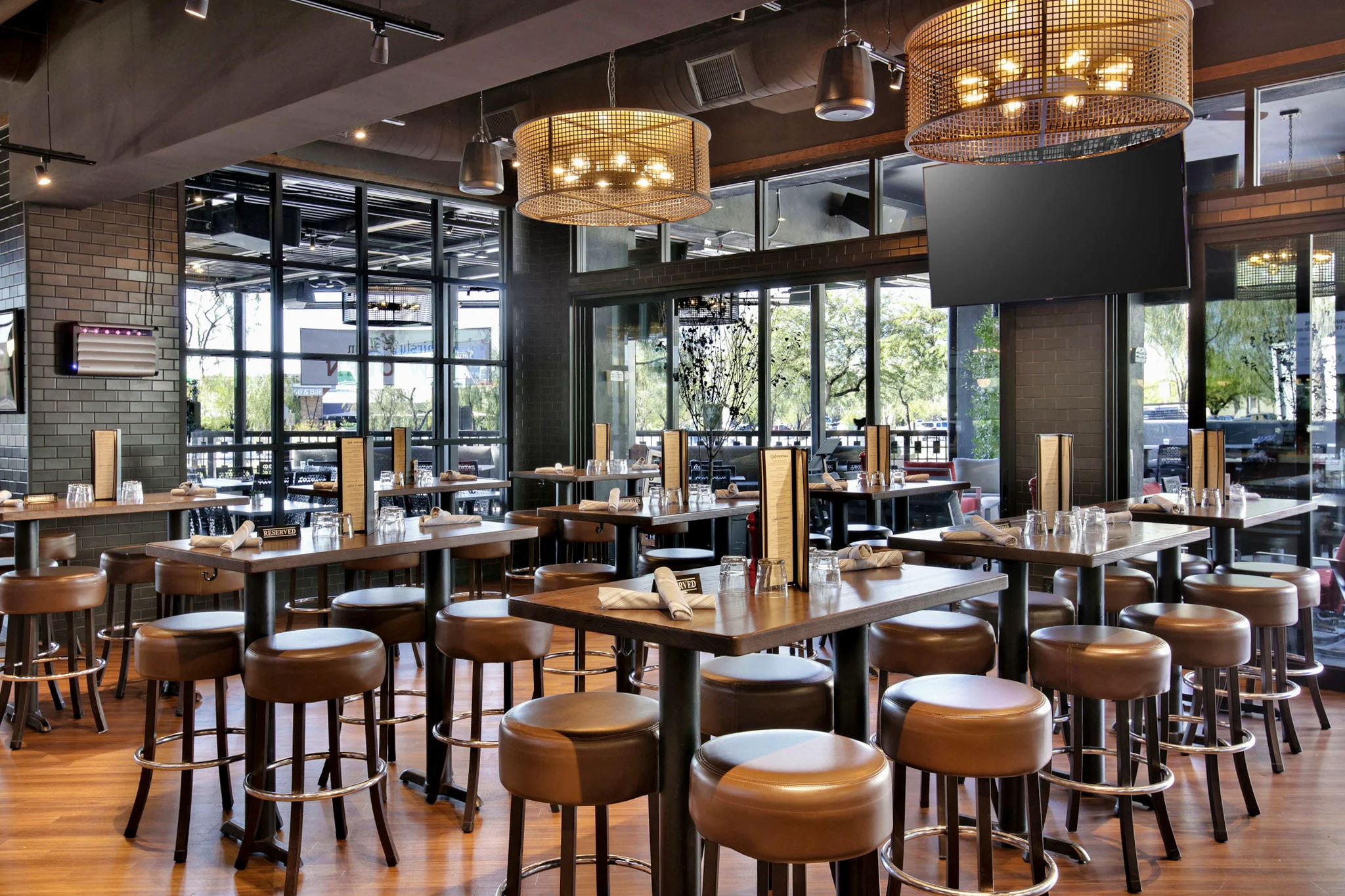 Interior of a restaurant with high-top tables, leather stools, and modern lighting, featuring large windows providing natural light.