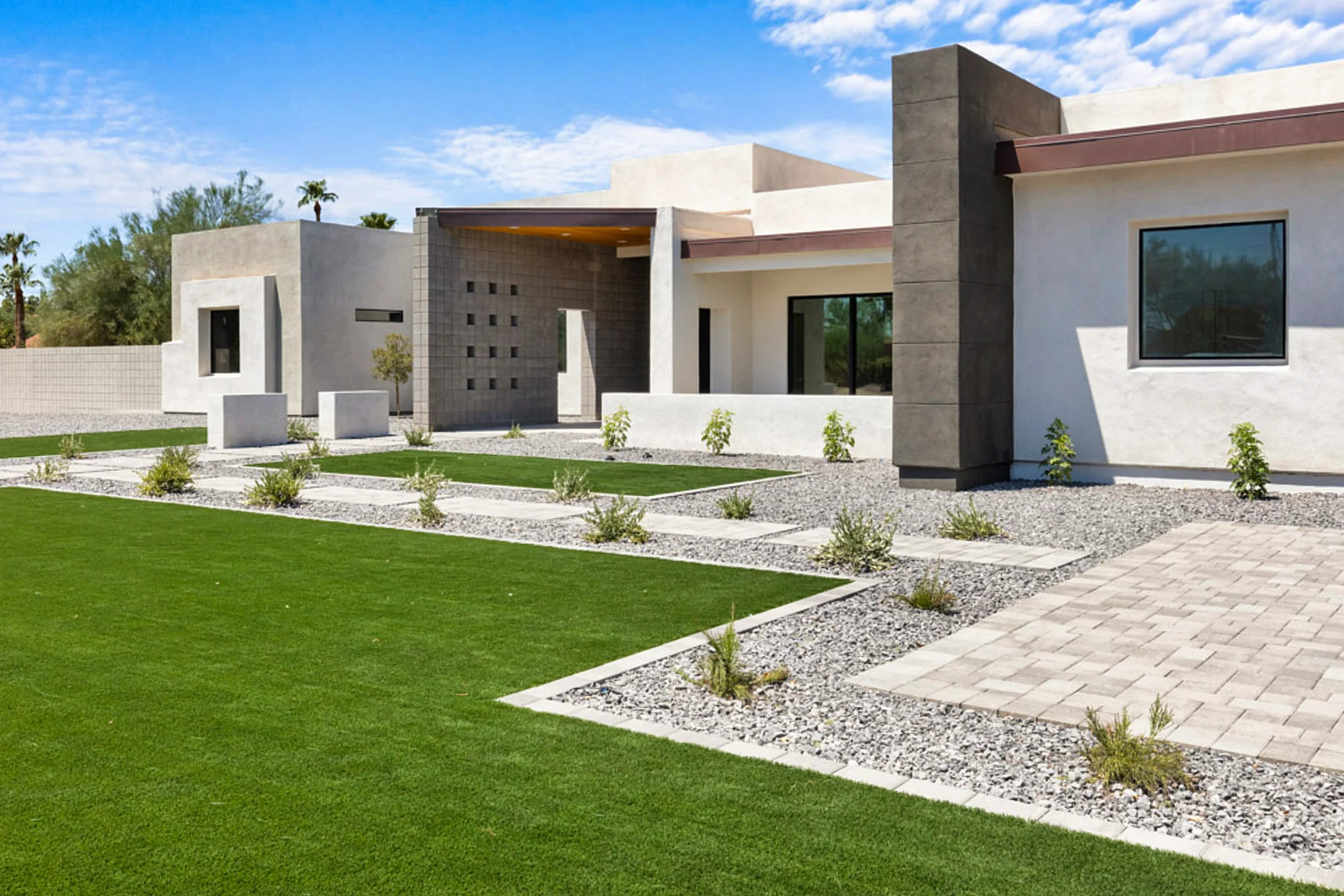 A modern desert home with clean architectural lines, landscaped with artificial grass, gravel, and minimalist plantings under a bright blue sky.