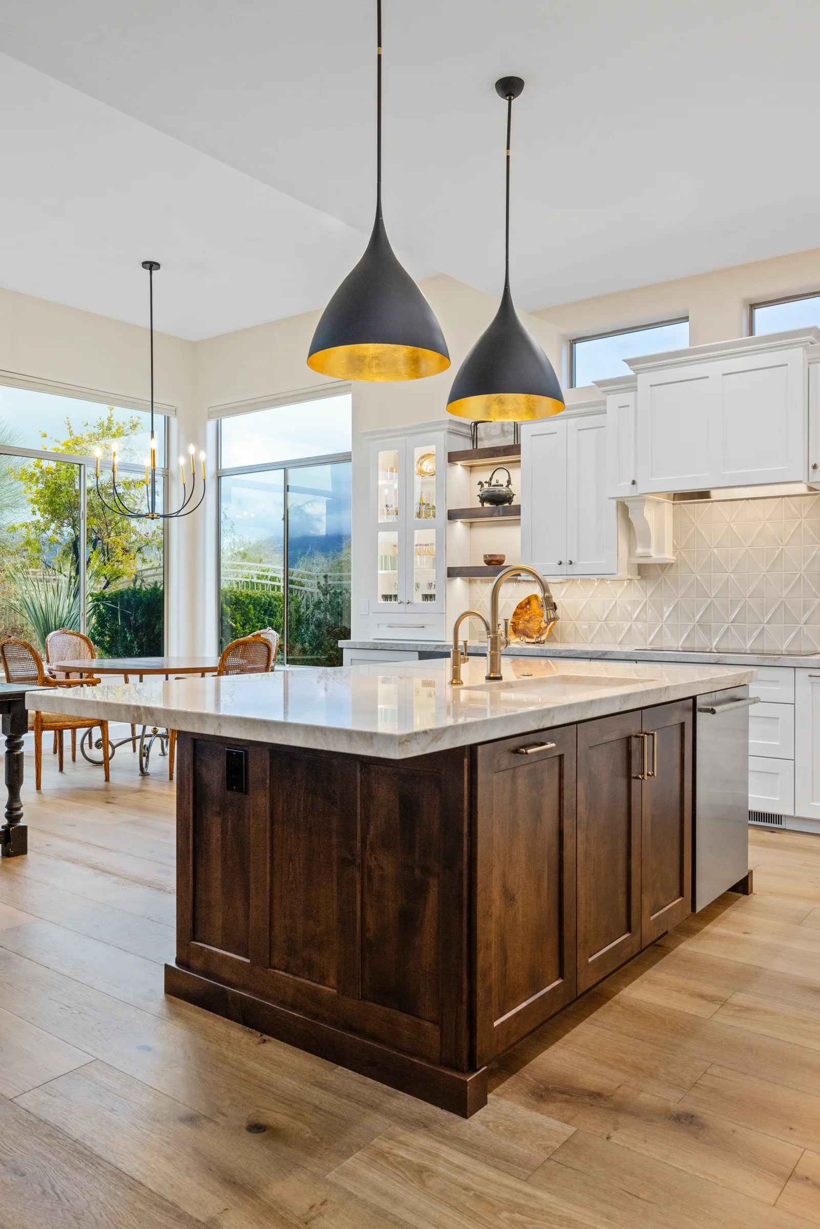 Modern kitchen with a large dark wood island, white cabinetry, gold-accented pendant lights, and a dining area with large windows offering natural light.