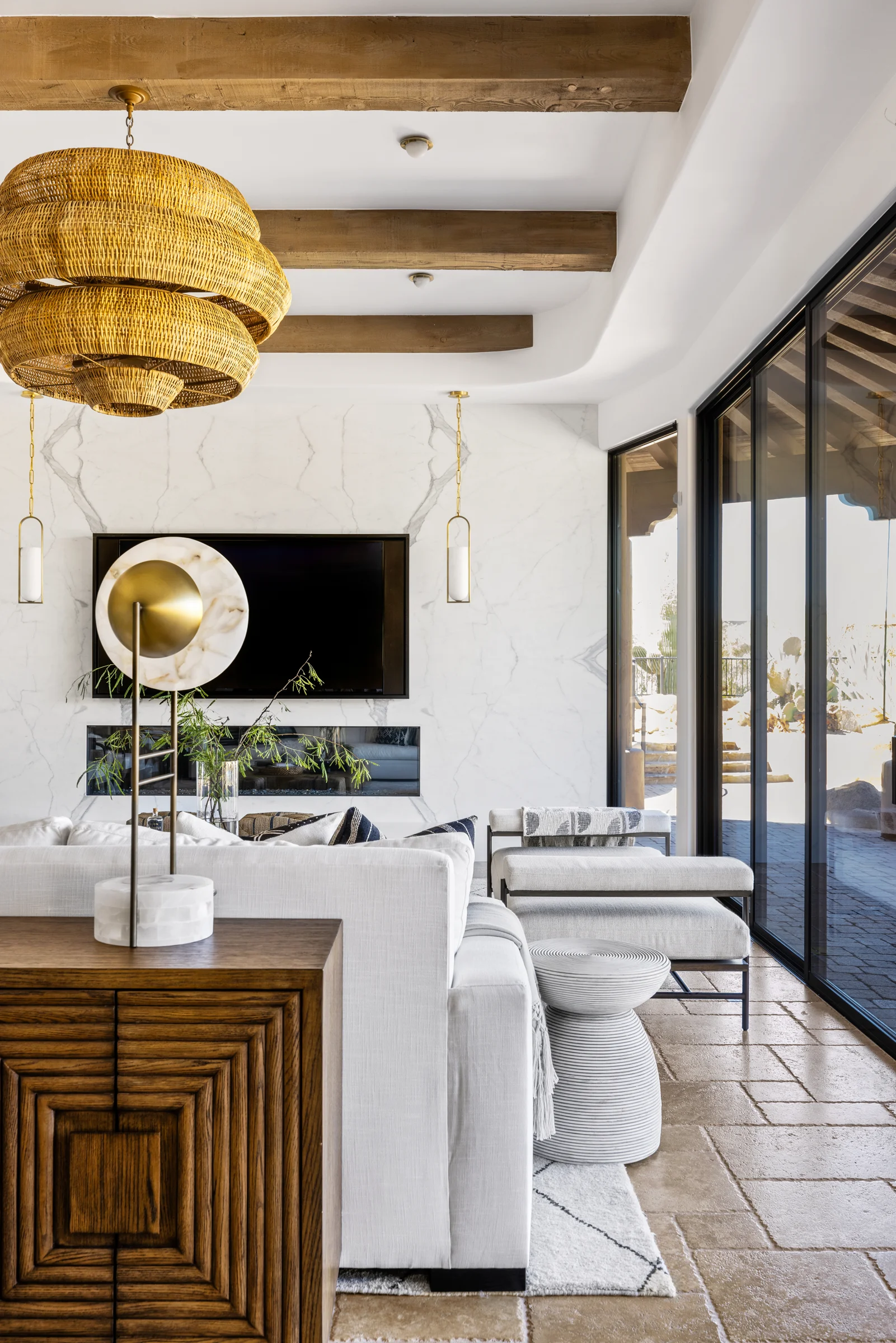 Modern living room with exposed wooden beams, a large marble fireplace wall, a woven pendant light, and white furniture overlooking a patio.