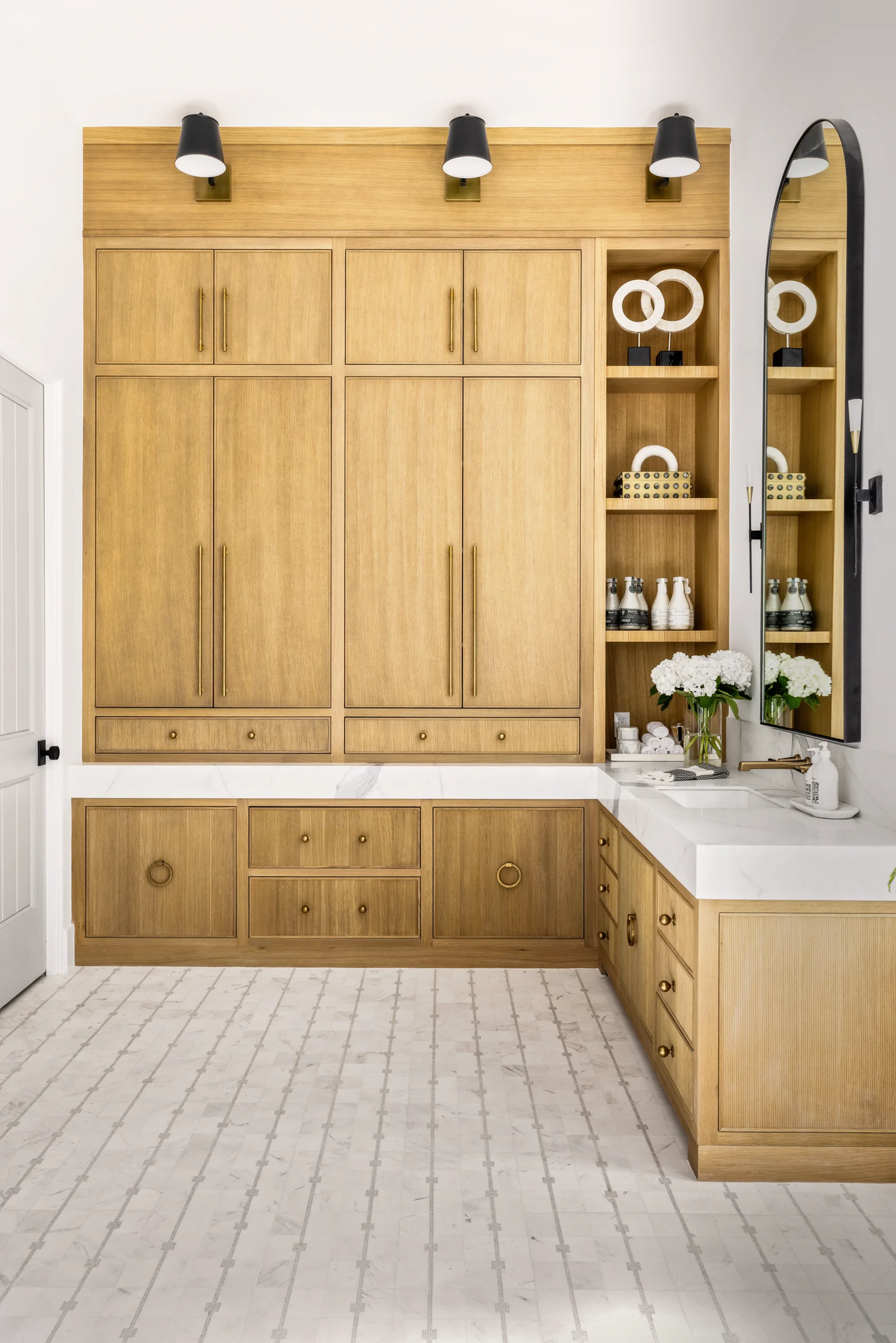 Luxurious bathroom with custom light wood cabinetry, a marble countertop, open shelving, and decorative accents, featuring a tall arched mirror.