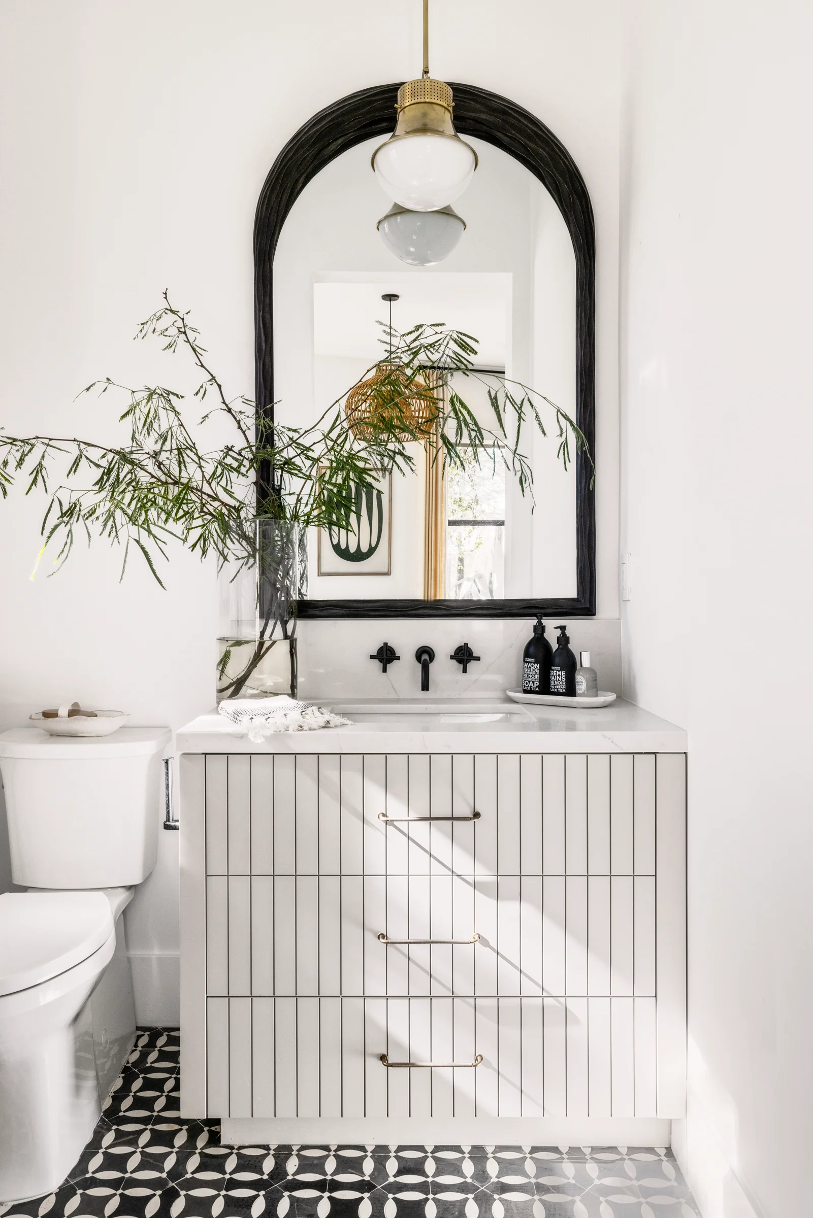 Bright bathroom with a white, vertically striped vanity, black hardware, arched mirror, and greenery in a glass vase.