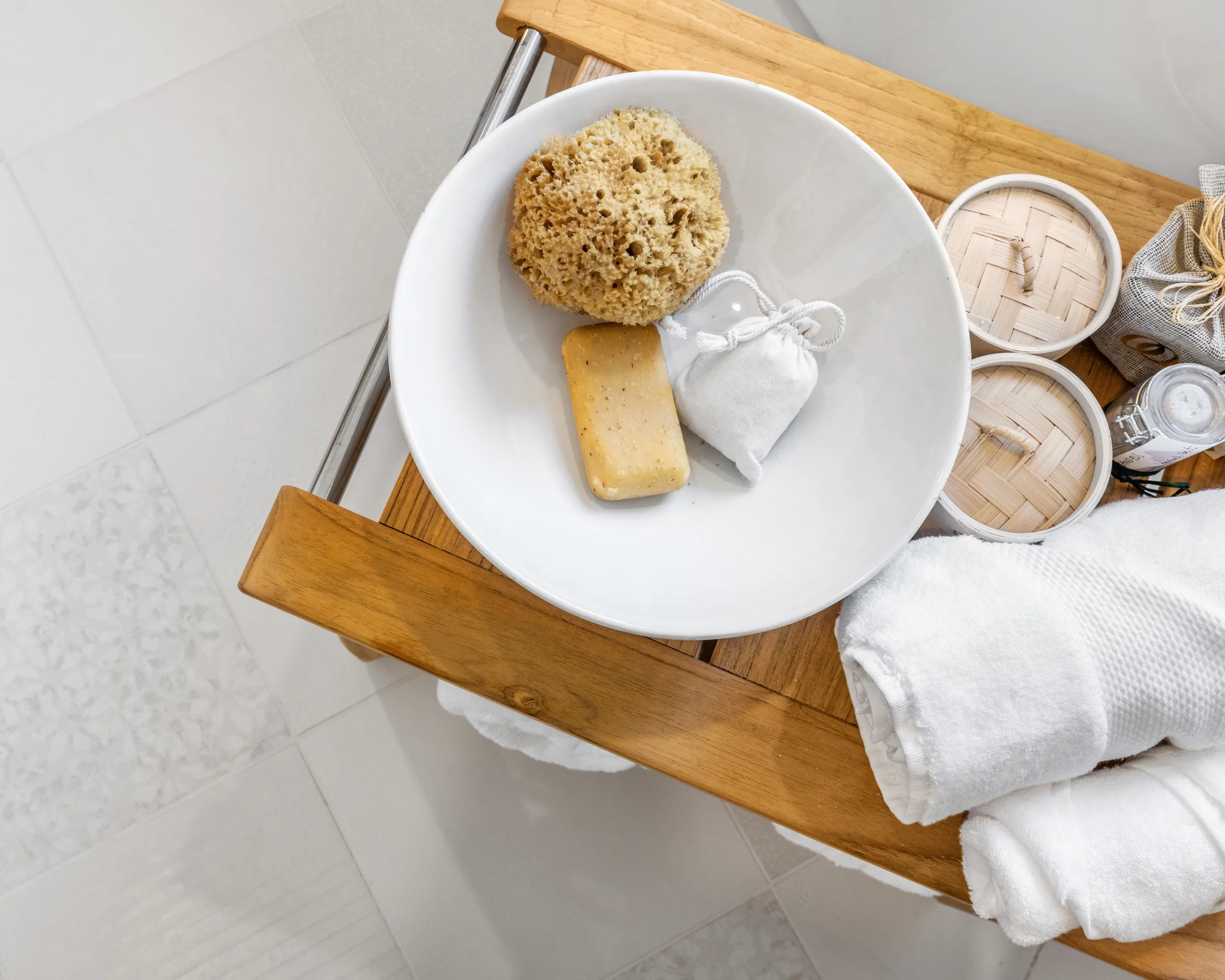 A wooden bath caddy with a white bowl holding a natural sponge, soap, and a sachet, alongside neatly rolled white towels and bamboo containers.